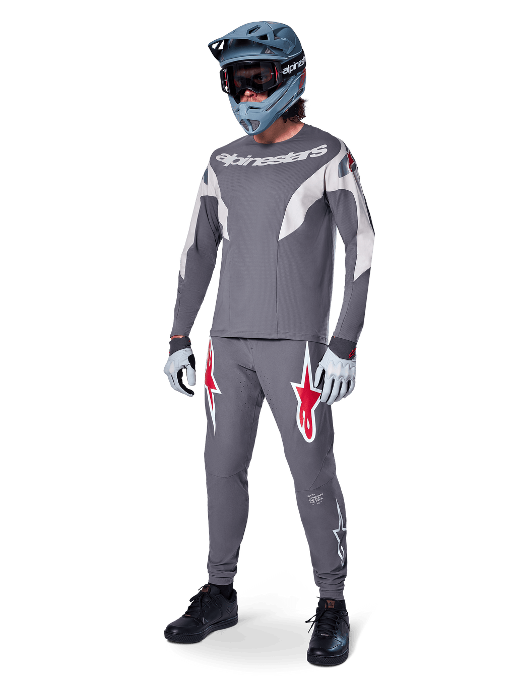 Alpinestars A-Supra Race Pants in Dark Gray, featuring bold red and white logos on the thighs, worn by a rider as part of a complete mountain bike racing kit with a matching gray jersey, blue-gray full-face helmet, and light gray gloves, high-performance gear for MTB racing