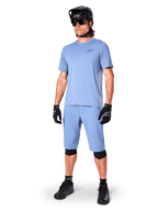 Modelo a usar calções Alpinestars A-Dura em Infinity Blue, camisola de manga curta azul-claro a condizer, capacete de BTT preto, óculos de sol e luvas pretas, equipamento completo de BTT para trail