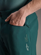 Calções Alpinestars A-Dura em Dark Pine, vista aproximada de uma mão no bolso com fecho e forro de rede, apresentando o logótipo da estrela Alpinestars branco e a marca A-Dura, construção têxtil durável para mountain biking