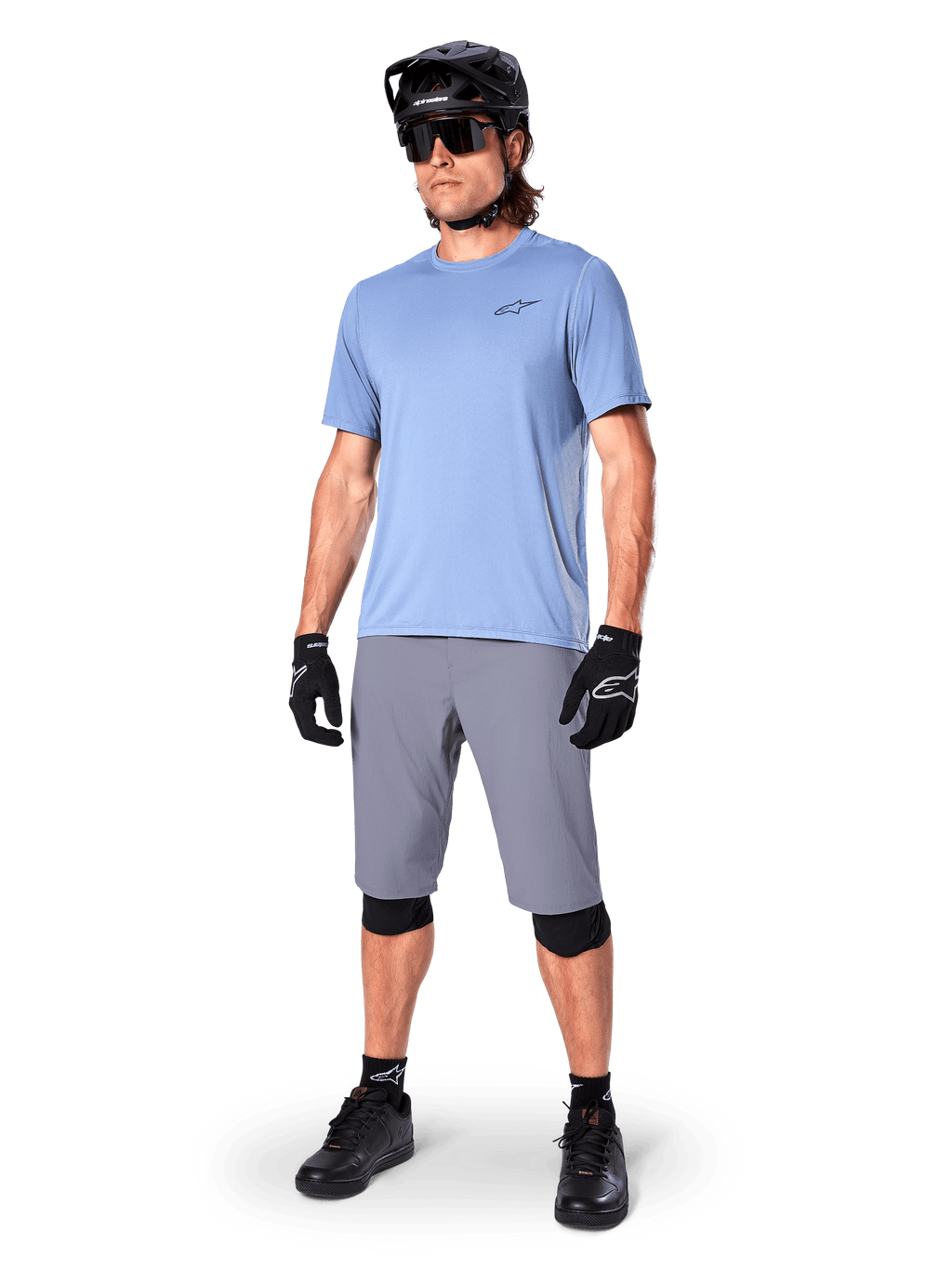 Calções Alpinestars A-Dura, calções de MTB, cinzento escuro, usados por um ciclista com uma camisola azul claro, capacete de MTB preto, luvas pretas e joelheiras, equipamento completo de mountain bike para trilhos e prática fora de estrada
