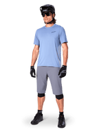 Calções Alpinestars A-Dura, calções de MTB, cinzento escuro, usados por um ciclista com uma camisola azul claro, capacete de MTB preto, luvas pretas e joelheiras, equipamento completo de mountain bike para trilhos e prática fora de estrada