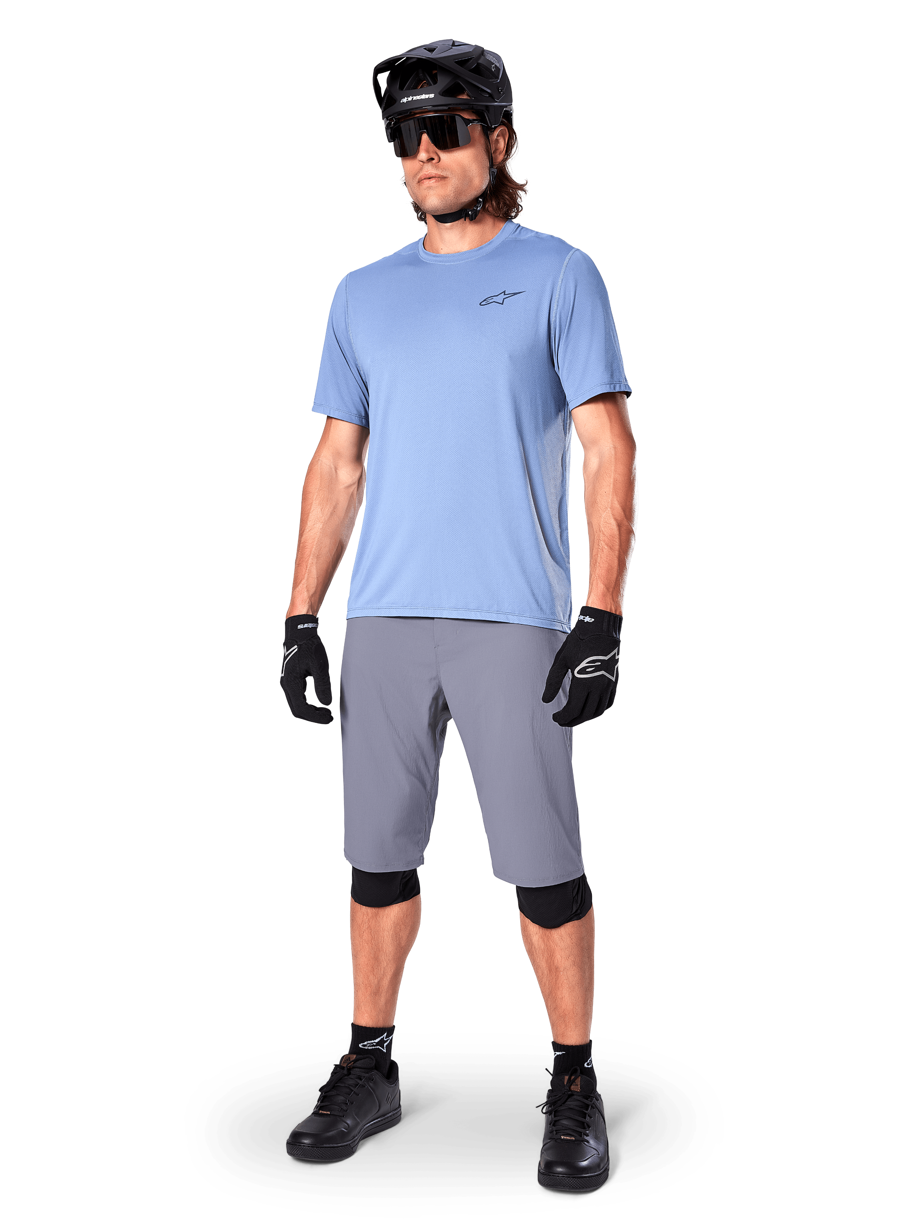 Calções Alpinestars A-Dura, calções de MTB, cinzento escuro, usados por um ciclista com uma camisola azul claro, capacete de MTB preto, luvas pretas e joelheiras, equipamento completo de mountain bike para trilhos e prática fora de estrada