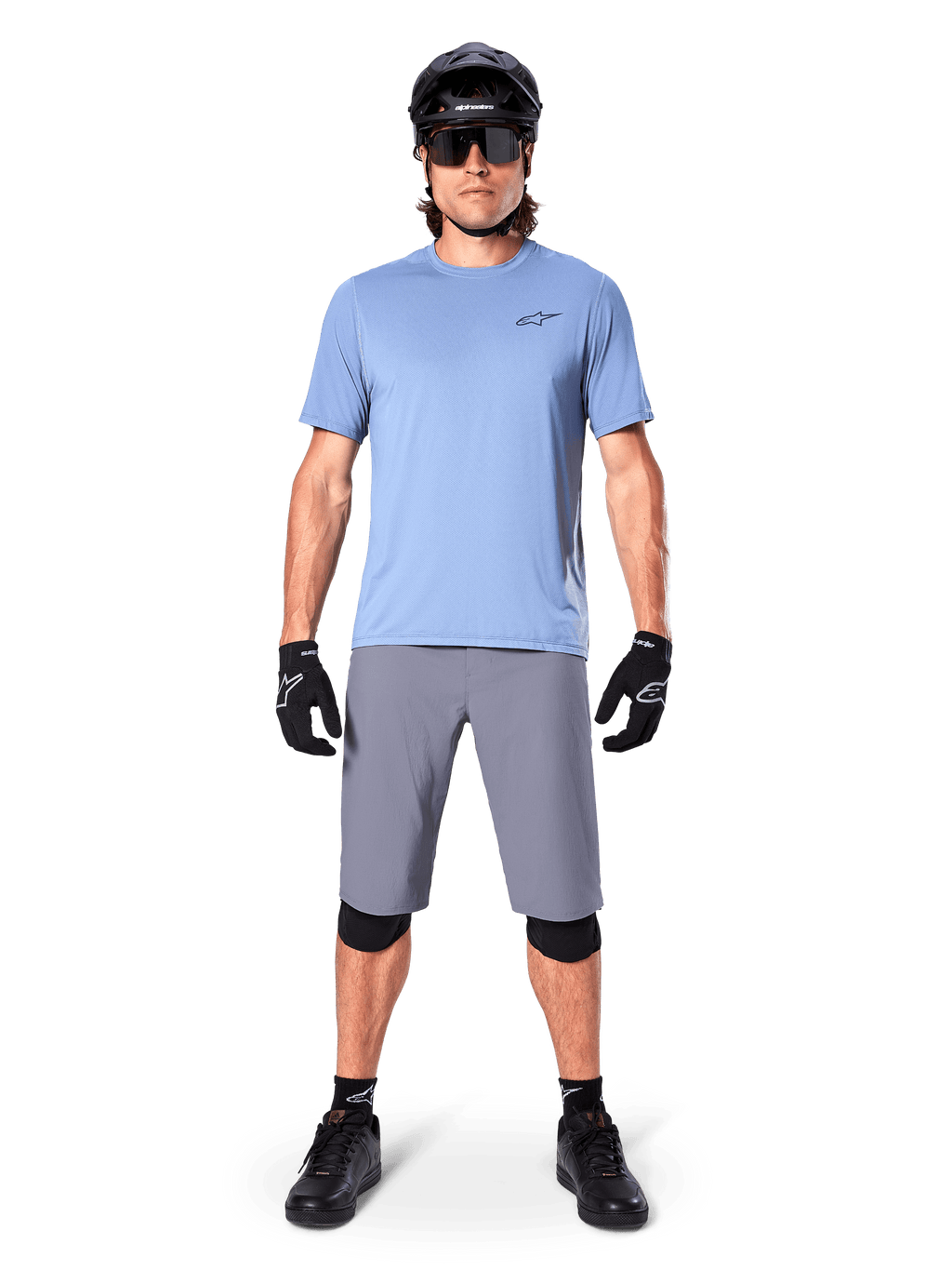 Ciclista masculino com calções Alpinestars A-Dura em cinzento escuro, combinados com uma t-shirt de manga curta azul-claro, capacete preto, óculos de sol e joelheiras pretas, equipamento completo de mountain biking para performance em todo-o-terreno.