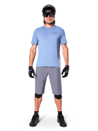 Ciclista masculino com calções Alpinestars A-Dura em cinzento escuro, combinados com uma t-shirt de manga curta azul-claro, capacete preto, óculos de sol e joelheiras pretas, equipamento completo de mountain biking para performance em todo-o-terreno.