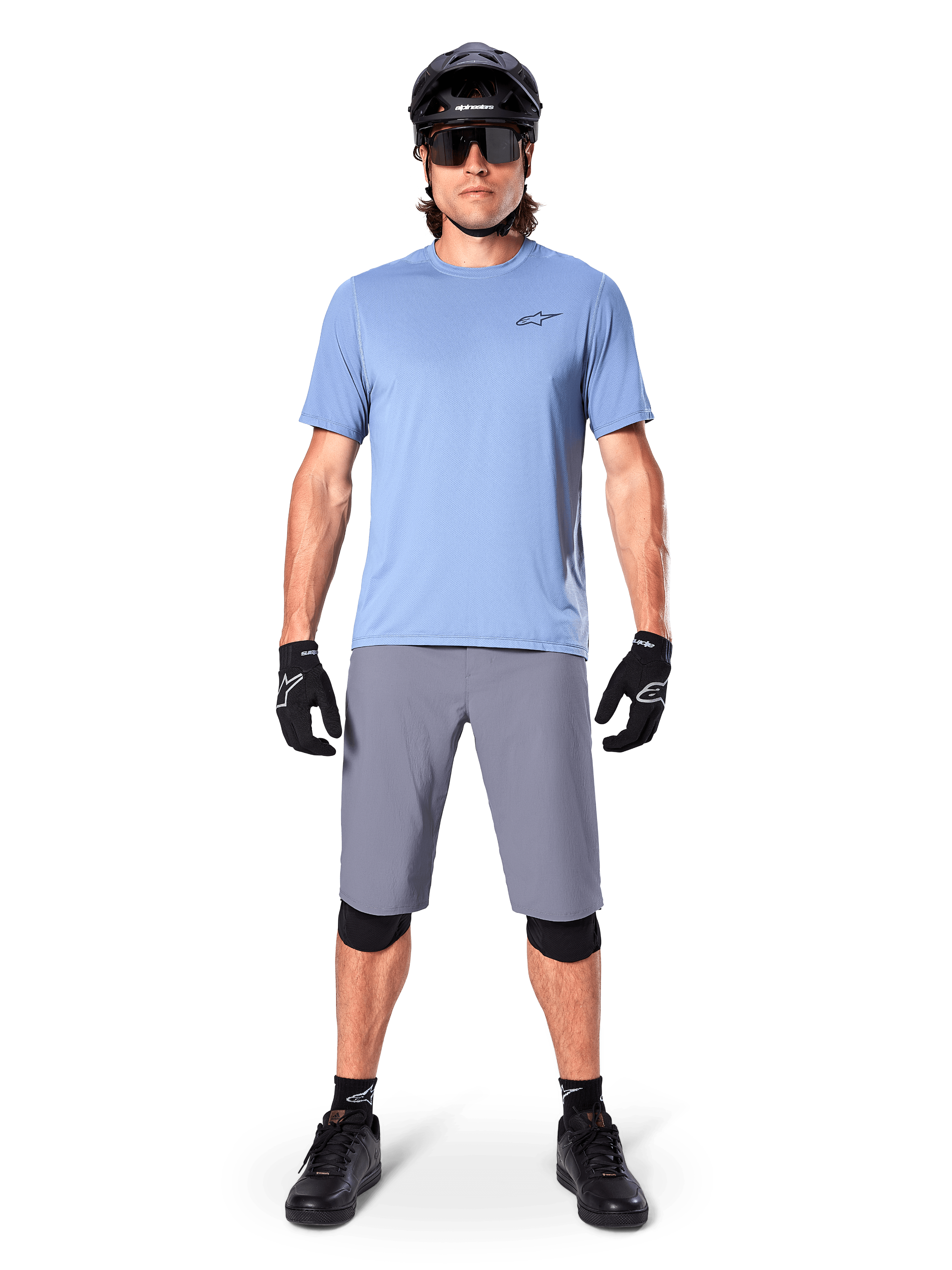 Ciclista masculino com calções Alpinestars A-Dura em cinzento escuro, combinados com uma t-shirt de manga curta azul-claro, capacete preto, óculos de sol e joelheiras pretas, equipamento completo de mountain biking para performance em todo-o-terreno.