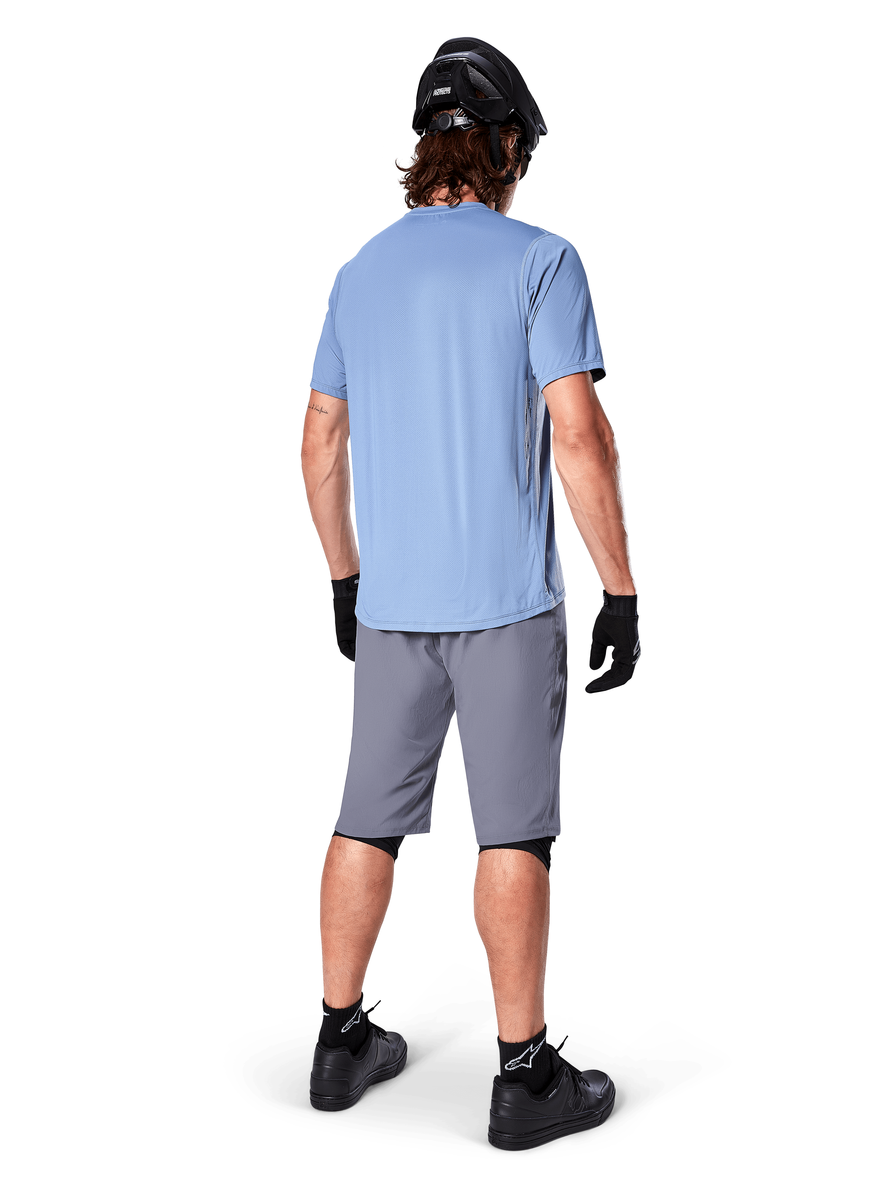 Calções Alpinestars A-Dura, calções de BTT cinzento escuro, vista traseira, usados com camisola de manga curta azul claro e capacete de BTT preto, luvas e meias pretas com sapatos pretos, equipamento funcional e durável para ciclismo todo-o-terreno