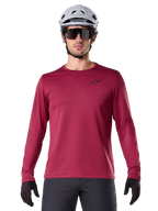 Ciclista masculino com Camisola Térmica Alpinestars A-Dura, camisola de BTT de manga comprida, cor bordô, combinada com capacete cinzento e óculos de sol pretos, apresenta um pequeno logótipo de estrela preto no peito, tecido de lã isolante para ciclismo de montanha em tempo frio