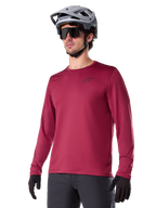 Alpinestars A-Dura Thermal Jersey, camisola de ciclismo de montanha de manga comprida, vermelho bordeaux, com forro de lã isolante e um pequeno logótipo preto no peito, usada com capacete cinzento, óculos de sol pretos e luvas pretas, concebida para ciclismo em tempo frio