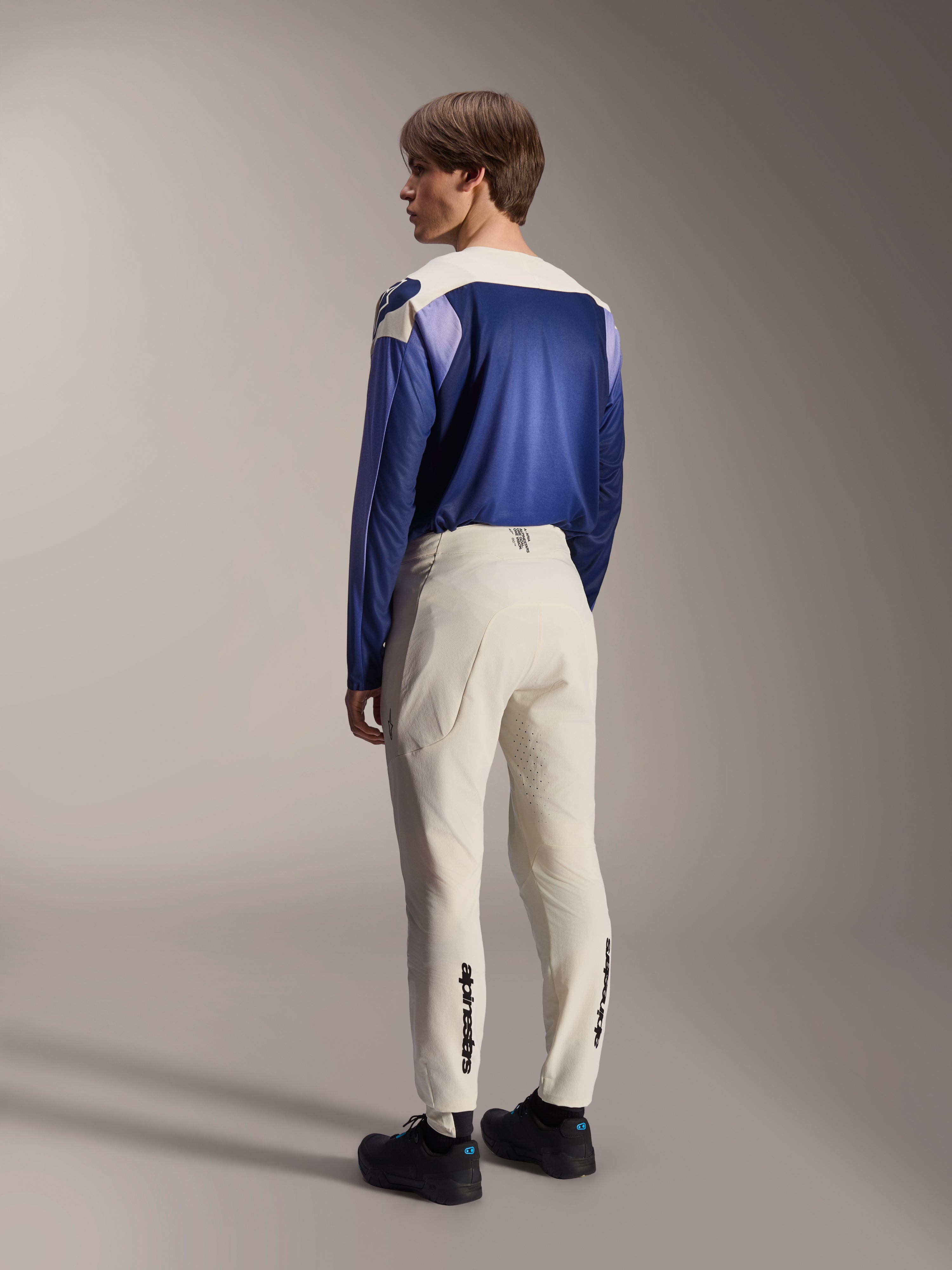 Alpinestars A-Aria Polartec Maker Elite Long Sleeve Jersey, camisola de manga comprida em Baja Blue, vista de costas num modelo masculino com calças de MTB em branco sujo e sapatos pretos, apresentando um design de blocos de cor em azul e branco, equipamento de performance para ciclismo de montanha