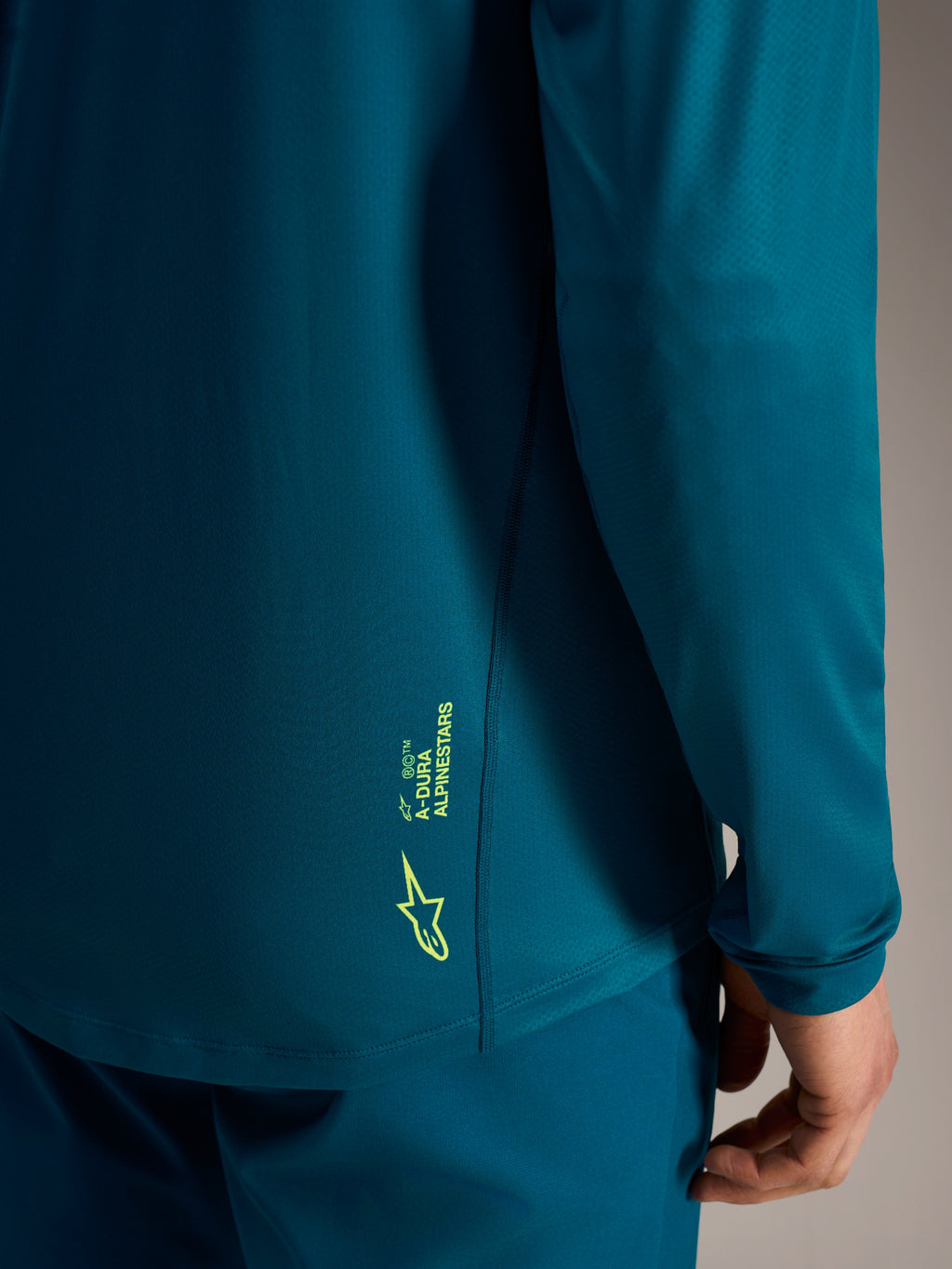 Camisola A-Dura Ride azul esverdeada com logótipo Alpinestars verde-lima na bainha inferior traseira texturizada.