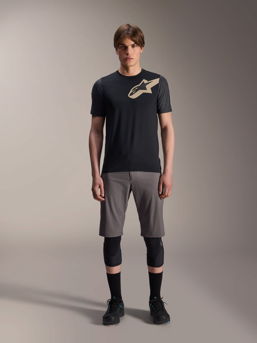 Alpinestars A-Dura Lipan Short Sleeve Jersey, preta com mangas em cinzento mesclado e logótipo Astars bege no peito, vestida por um modelo masculino com calções de BTT cinzentos e joelheiras pretas, equipamento de performance para MTB