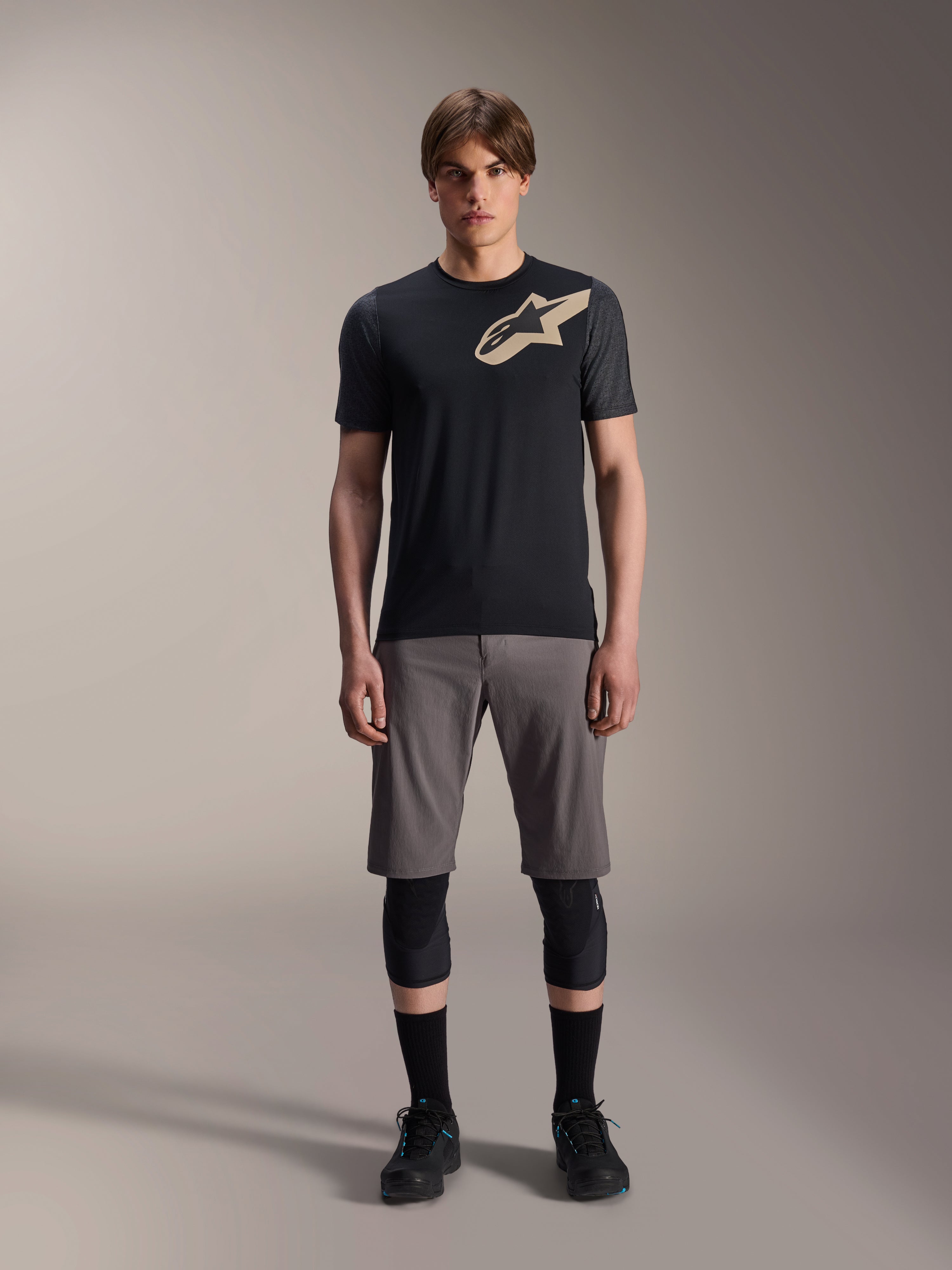 Alpinestars A-Dura Lipan Short Sleeve Jersey, preta com mangas em cinzento mesclado e logótipo Astars bege no peito, vestida por um modelo masculino com calções de BTT cinzentos e joelheiras pretas, equipamento de performance para MTB