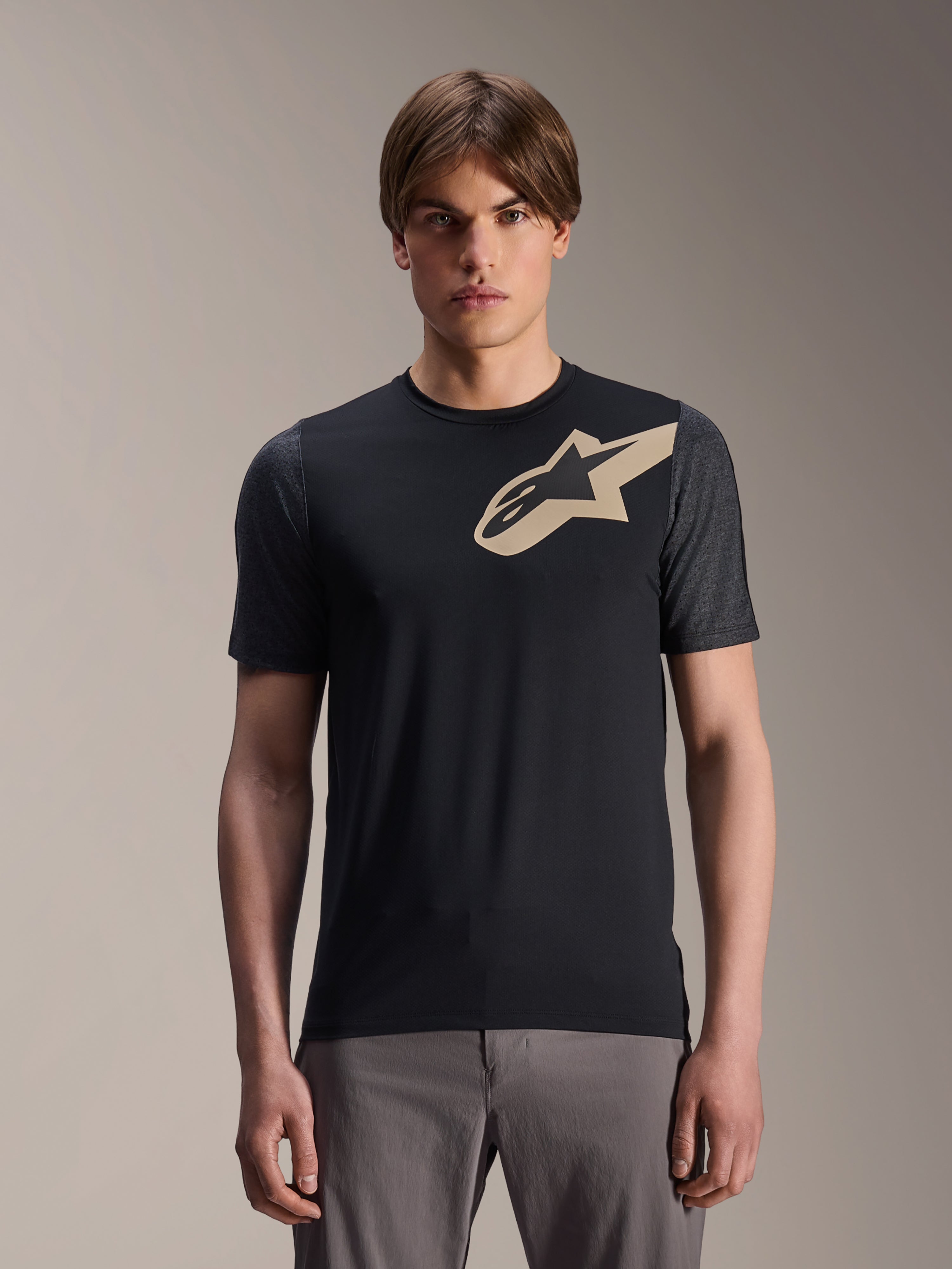 Modelo masculino a usar Jersey de Manga Curta Alpinestars A-Dura Lipan, camisola de BTT preta com logótipo de estrela bege e mangas cinzentas escuras, combinada com calças de ciclismo cinzentas, vestuário de performance casual