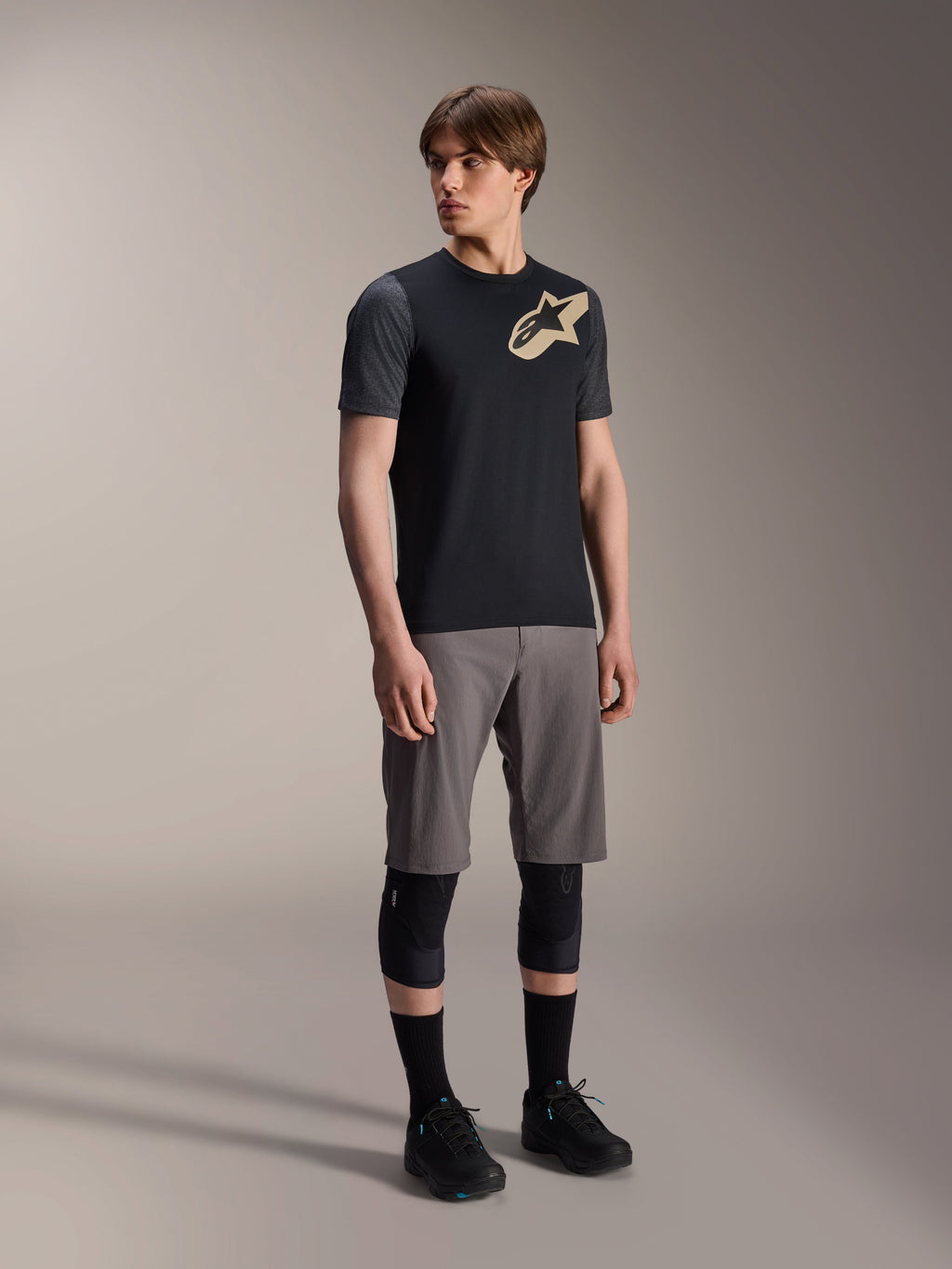 Modelo masculino a usar a Camisola de Manga Curta Alpinestars A-Dura Lipan em preto, com mangas cinzento-escuro e logótipo Astars bege no peito, combinada com calções de BTT cinzentos, joelheiras pretas e sapatilhas de ciclismo pretas, equipamento de BTT coordenado