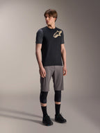 Modelo masculino a usar a Camisola de Manga Curta Alpinestars A-Dura Lipan em preto, com mangas cinzento-escuro e logótipo Astars bege no peito, combinada com calções de BTT cinzentos, joelheiras pretas e sapatilhas de ciclismo pretas, equipamento de BTT coordenado