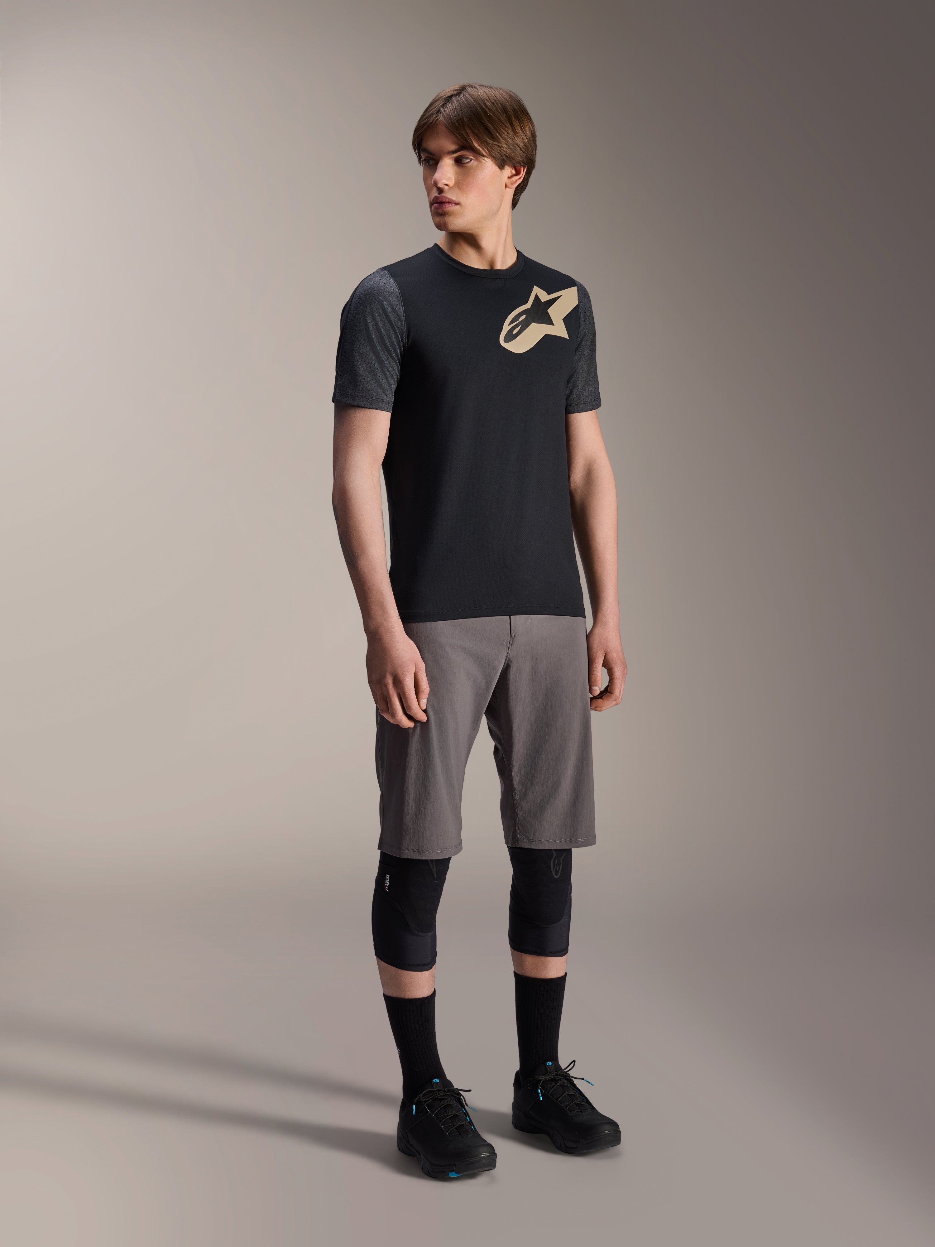 Modelo masculino a usar a Camisola de Manga Curta Alpinestars A-Dura Lipan em preto, com mangas cinzento-escuro e logótipo Astars bege no peito, combinada com calções de BTT cinzentos, joelheiras pretas e sapatilhas de ciclismo pretas, equipamento de BTT coordenado