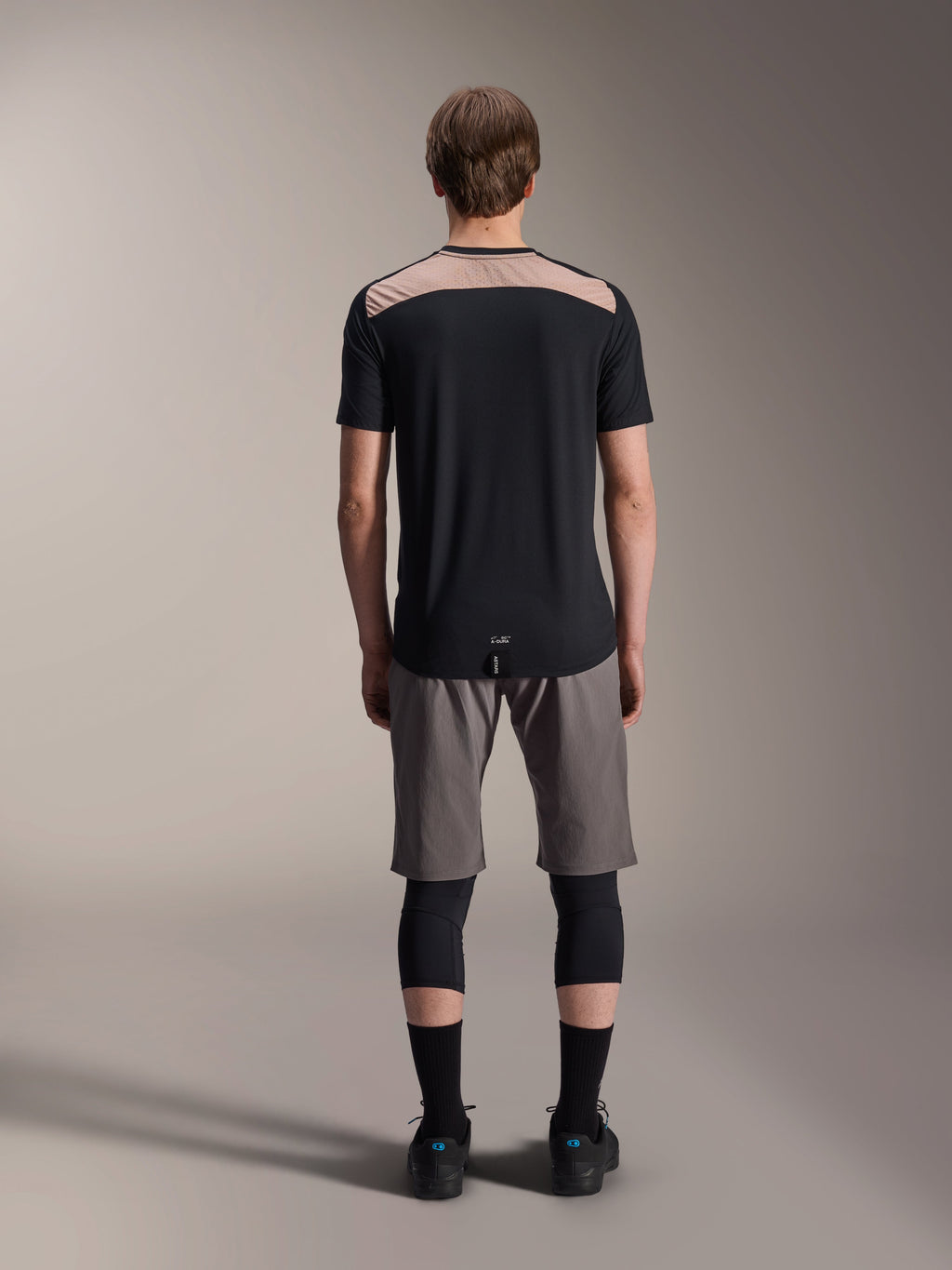 Camisola Alpinestars A-Dura Lipan - Manga Curta, preta com painel superior traseiro bege, vista traseira mostrando a cauda mais longa, usada com calções de BTT cinzentos e joelheiras pretas, vestuário de performance para bicicleta de montanha
