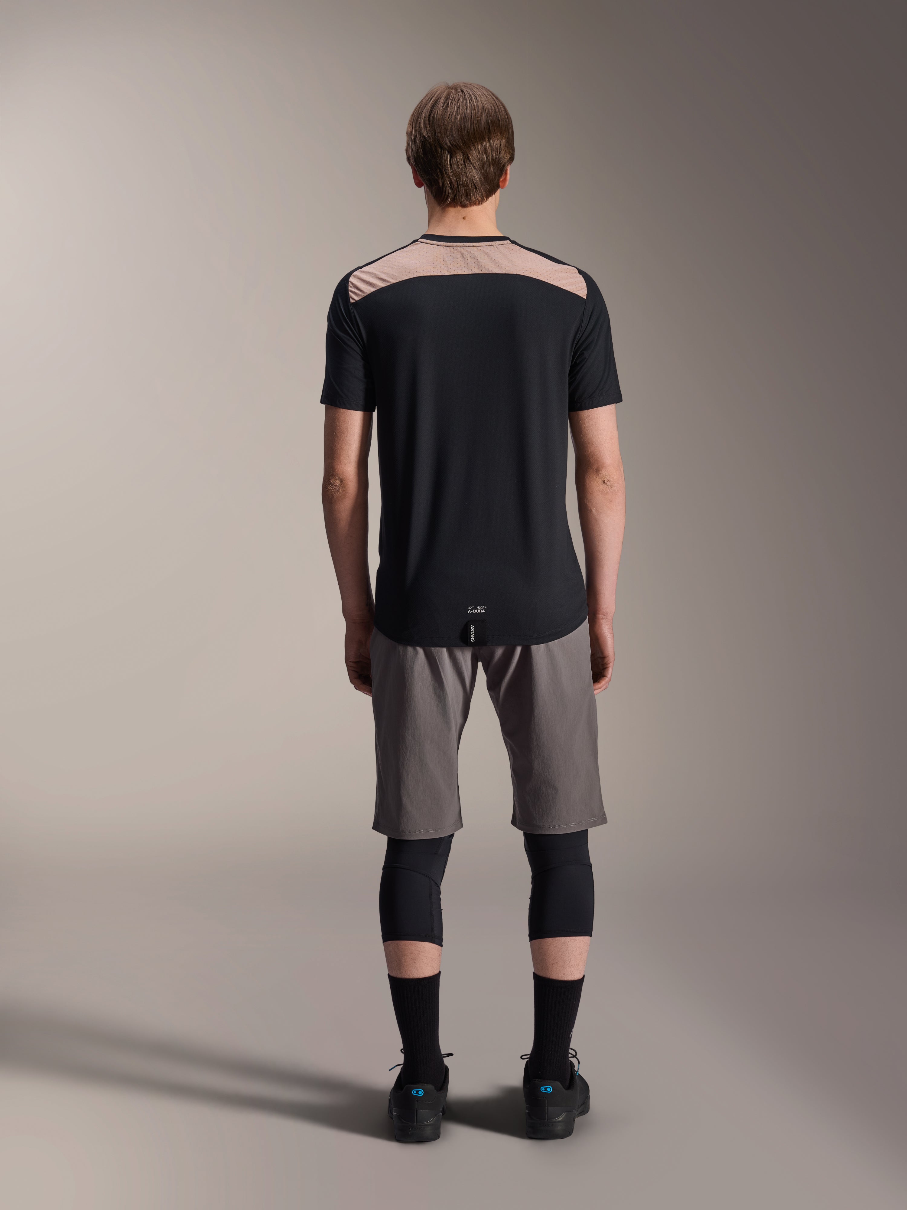 Camisola Alpinestars A-Dura Lipan - Manga Curta, preta com painel superior traseiro bege, vista traseira mostrando a cauda mais longa, usada com calções de BTT cinzentos e joelheiras pretas, vestuário de performance para bicicleta de montanha