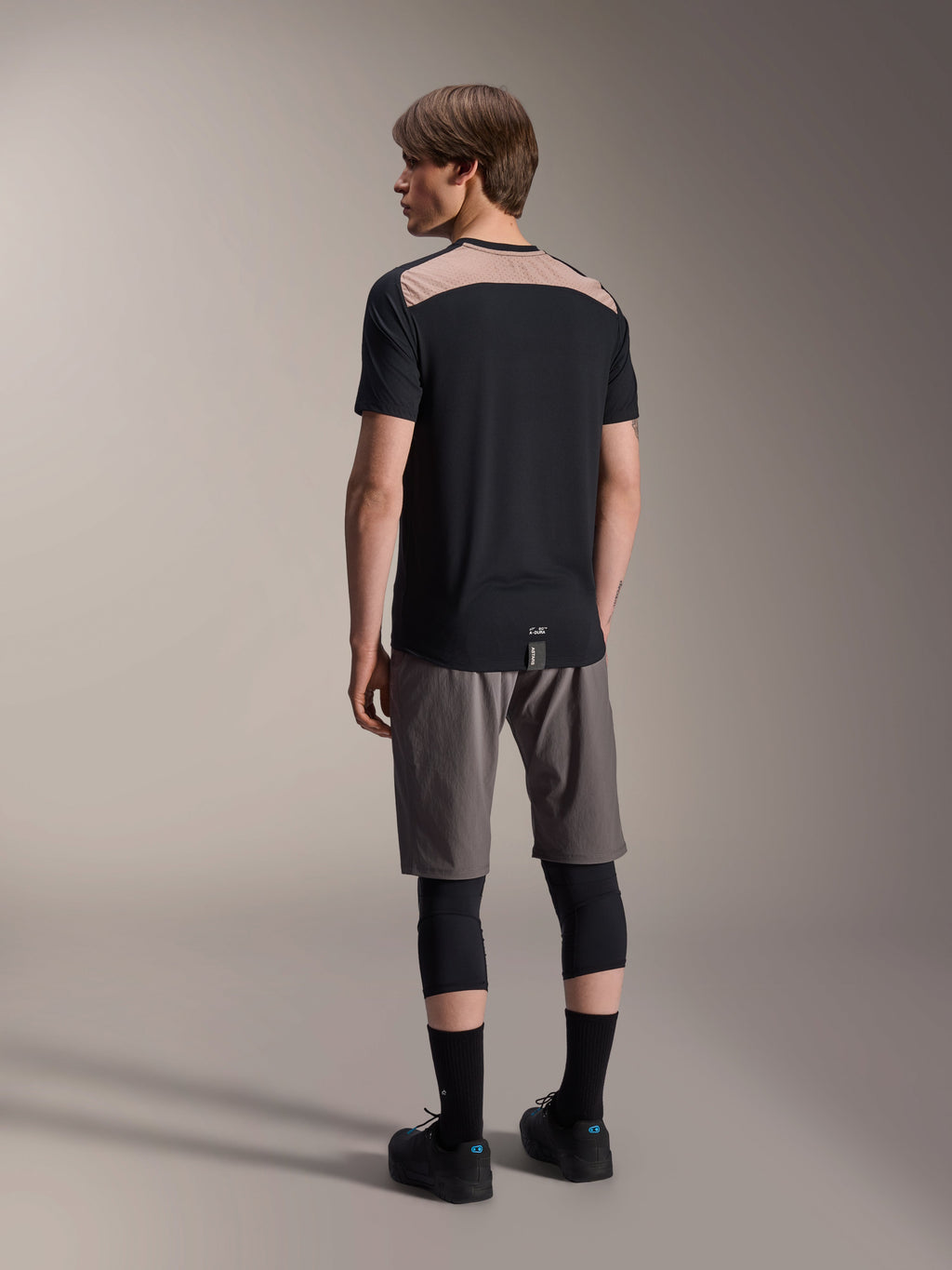 Alpinestars A-Dura Lipan Jersey de Manga Curta em preto, vista traseira no modelo a destacar o painel de malha perfurada castanho claro na parte superior das costas, combinada com calções de BTT cinzentos e joelheiras pretas, conjunto de trail completo.