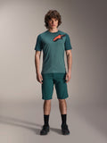 Calções Alpinestars A-Dura em Dark Pine, calções de ciclismo de montanha para homem, apresentados num modelo com uma camisola de manga curta verde-azulado a condizer com logótipo de estrela vermelho e preto, meias pretas e sapatos de ciclismo, vestuário de ciclismo de montanha casual e desportivo