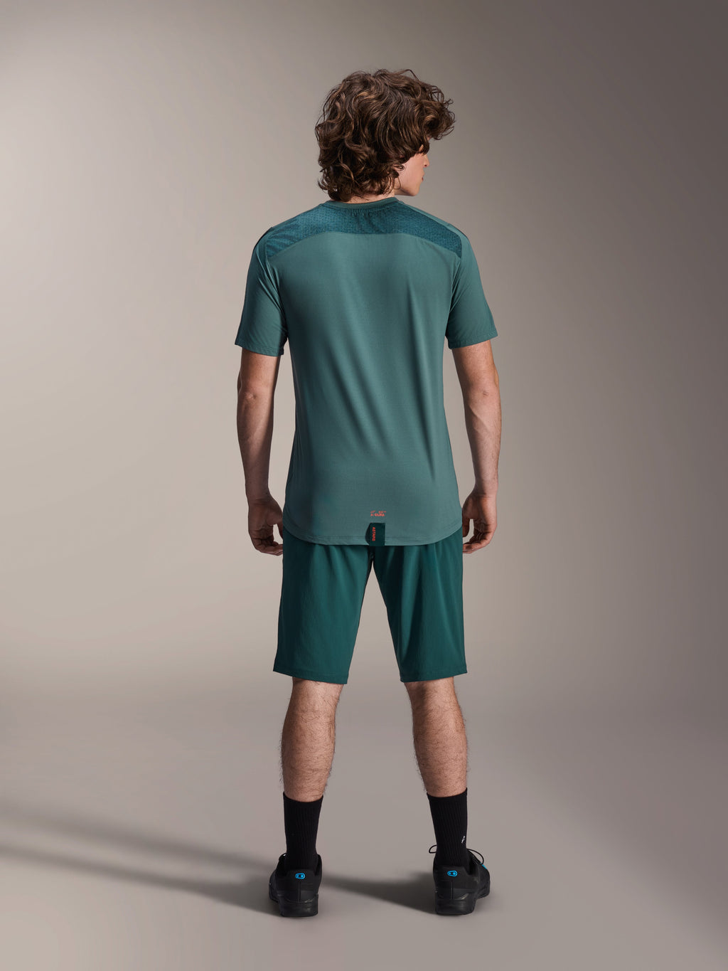 Camisola de manga curta Alpinestars A-Dura Lipan em Silver Pine, vista traseira a mostrar os painéis de jacquard texturizados nos ombros e a bainha com cauda mais longa, usada com calções de bicicleta de montanha em verde escuro a condizer e calçado preto.