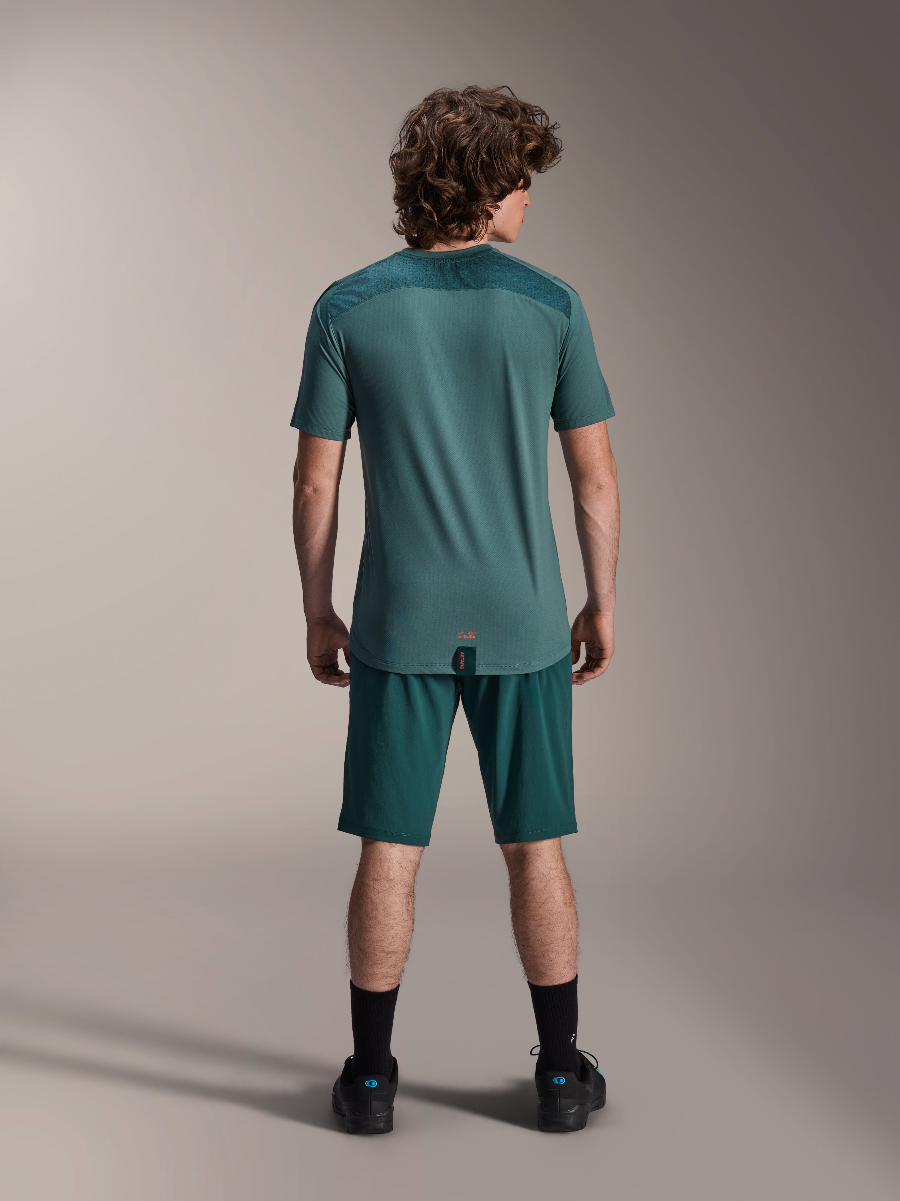 Camisola de manga curta Alpinestars A-Dura Lipan em Silver Pine, vista traseira a mostrar os painéis de jacquard texturizados nos ombros e a bainha com cauda mais longa, usada com calções de bicicleta de montanha em verde escuro a condizer e calçado preto.