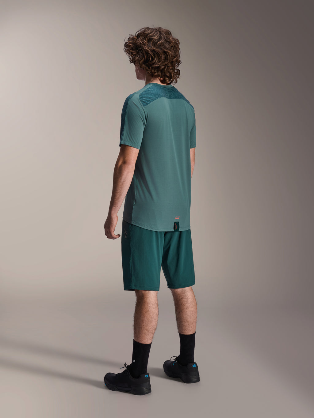 Modelo a usar calções Alpinestars A-Dura em Dark Pine e camisola de manga curta Lipan verde a condizer, vista traseira, combinados com meias pretas e sapatos de BTT pretos, equipamento profissional de ciclismo de montanha
