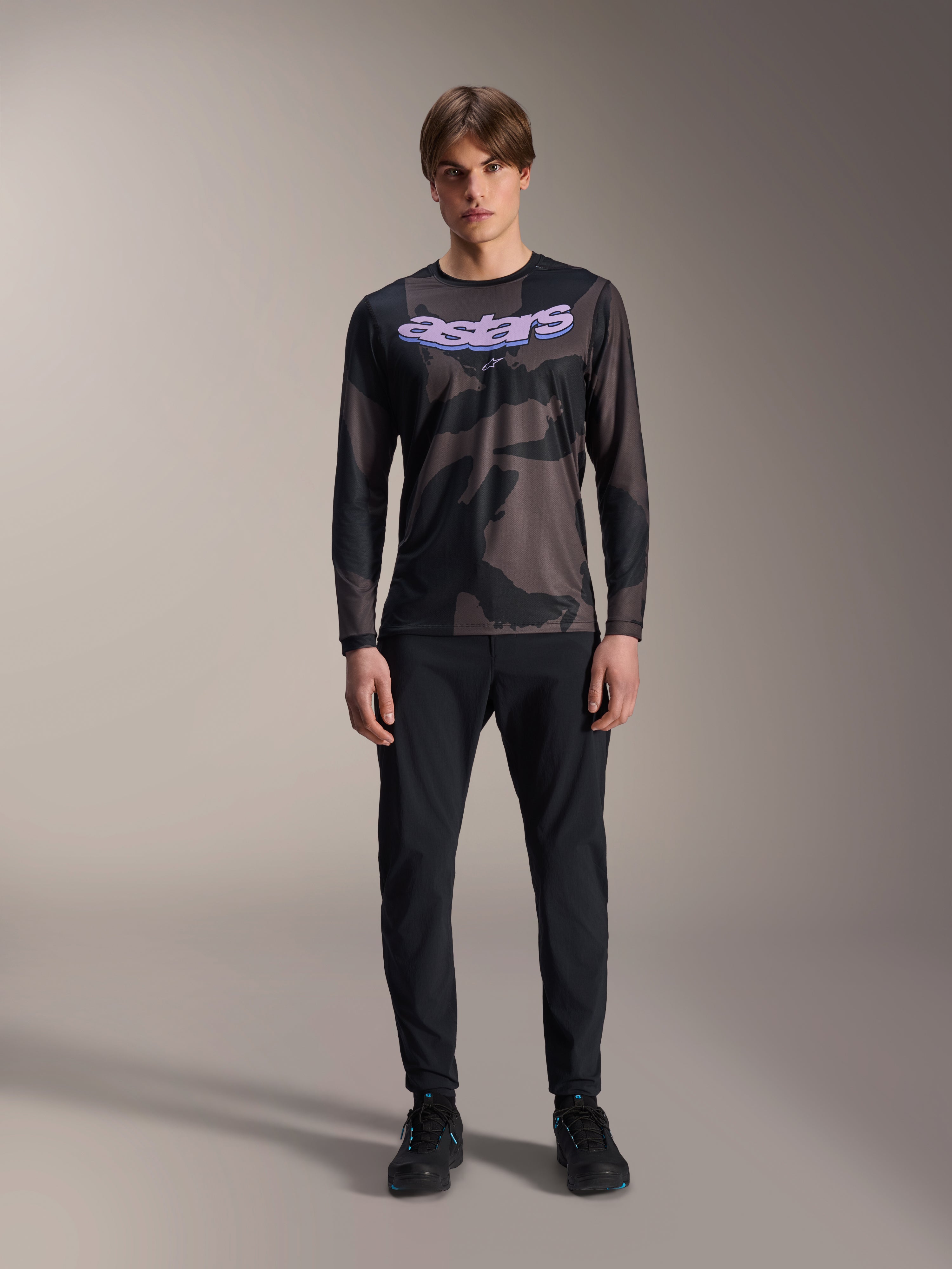 Modelo a vestir camisola de manga comprida Alpinestars A-Dura Hollow Camo em Black Camo, com logótipo 'astars' roxo no peito, combinada com calças pretas e sapatos de ciclismo pretos, equipamento de ciclismo de montanha