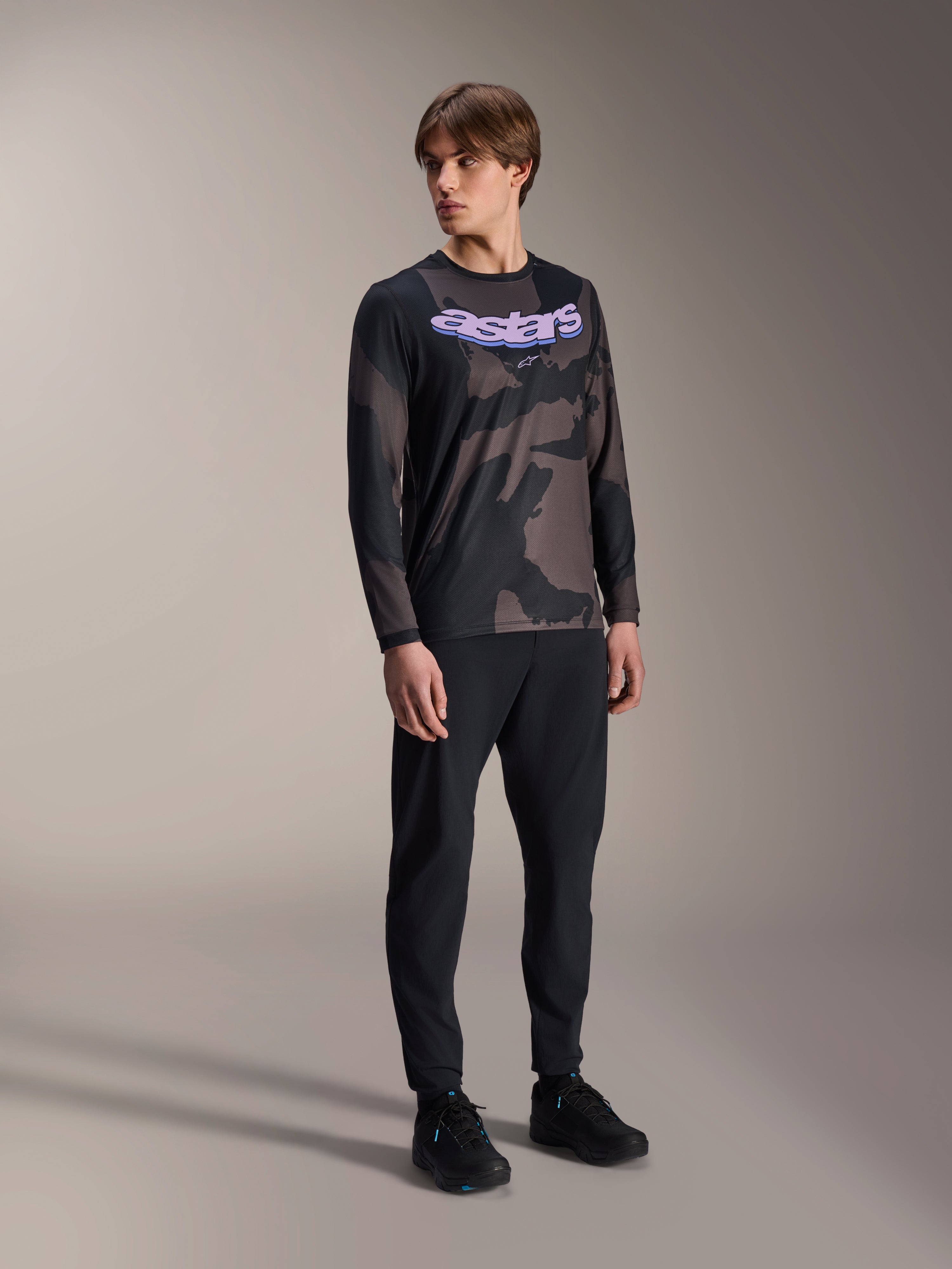Modelo masculino a usar a Camisola de Manga Comprida Alpinestars A-Dura Hollow Camo, padrão Black Camo com gráfico 'astars' roxo no peito, combinada com calças técnicas pretas e sapatos pretos, equipamento casual de BTT