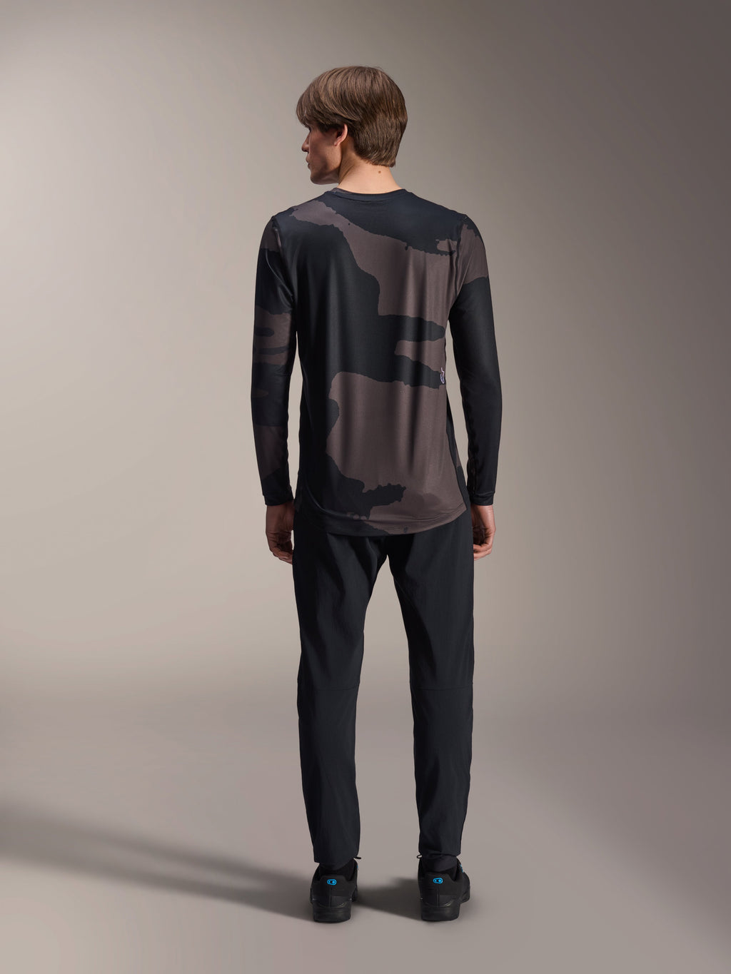 Alpinestars A-Dura Hollow Camo Long Sleeve Jersey, Black Camo, vista traseira vestida com calças pretas, com um padrão de camuflagem escuro e bainha traseira alongada para cobertura em BTT.