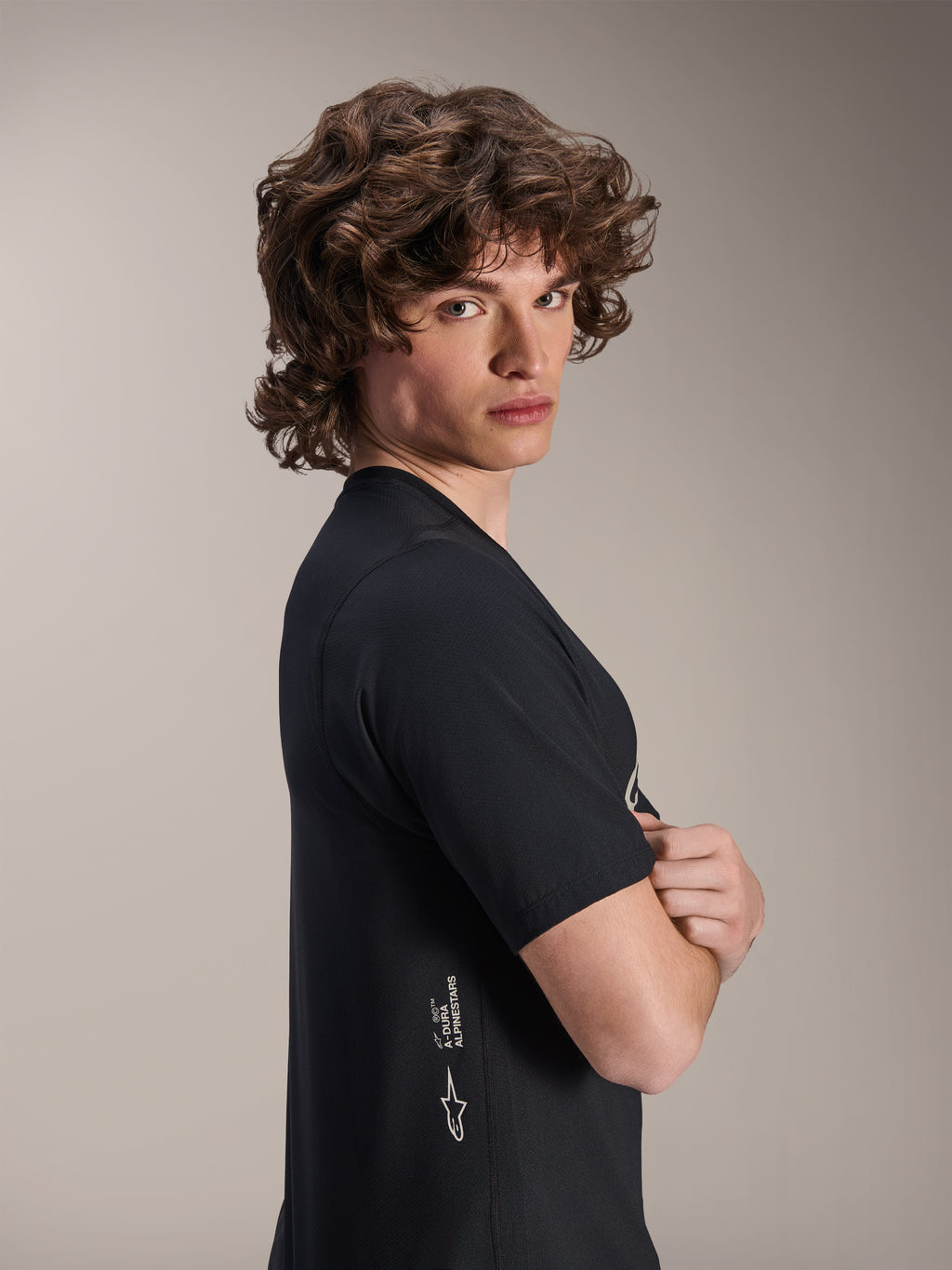 Alpinestars A-Dura Astar Jersey, camisola de BTT de manga curta, preta, modelo masculino posando de perfil com os braços cruzados, com logótipo Alpinestars a branco na parte lateral inferior, design atlético casual para ciclismo de montanha.
