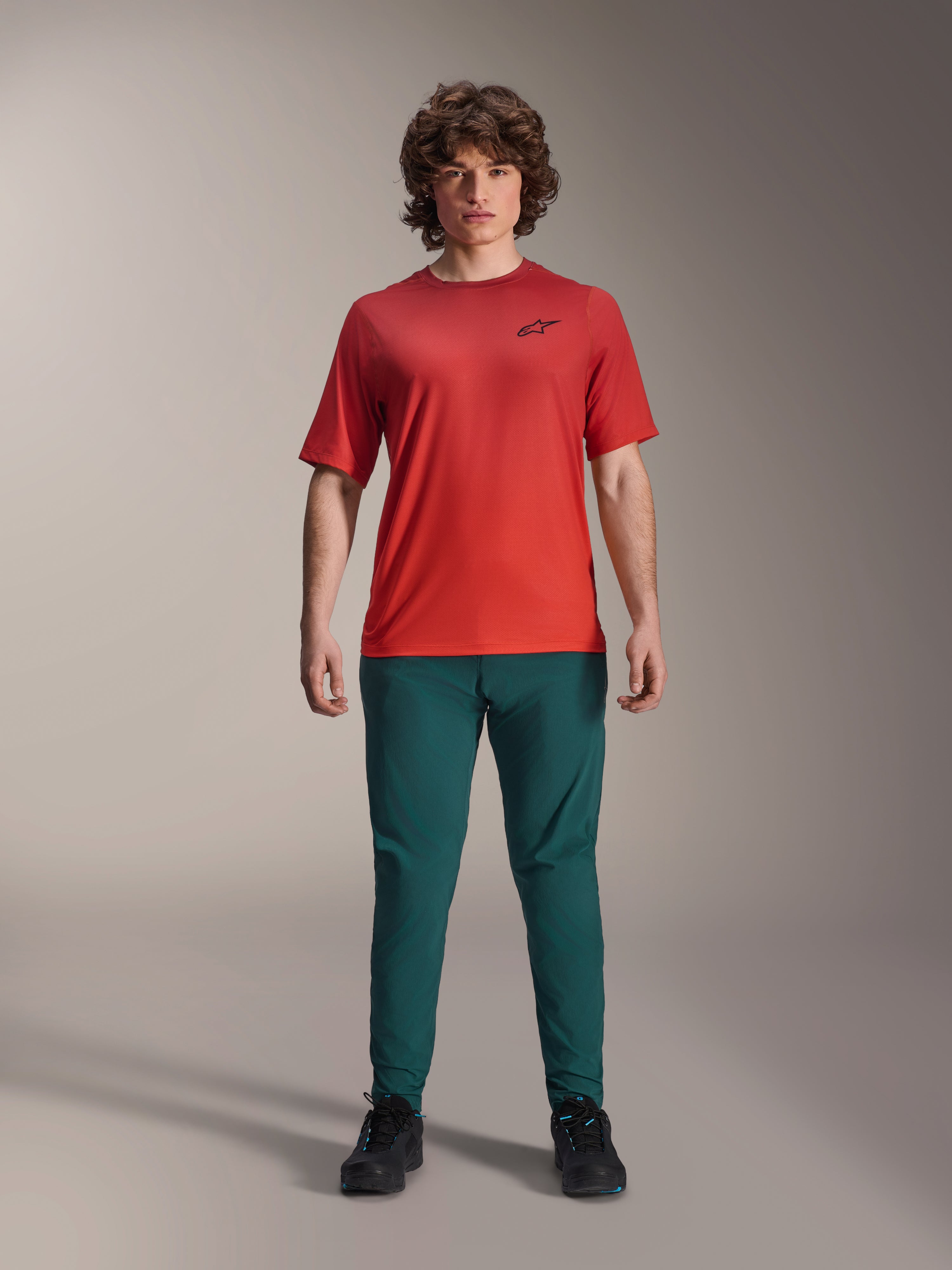 Calças Alpinestars A-Dura em verde pinho escuro combinadas com uma camisola de manga curta A-Dura Rise vermelha, modelo a posar com equipamento completo de ciclismo de montanha, ajuste de performance casual, vestuário de BTT durável