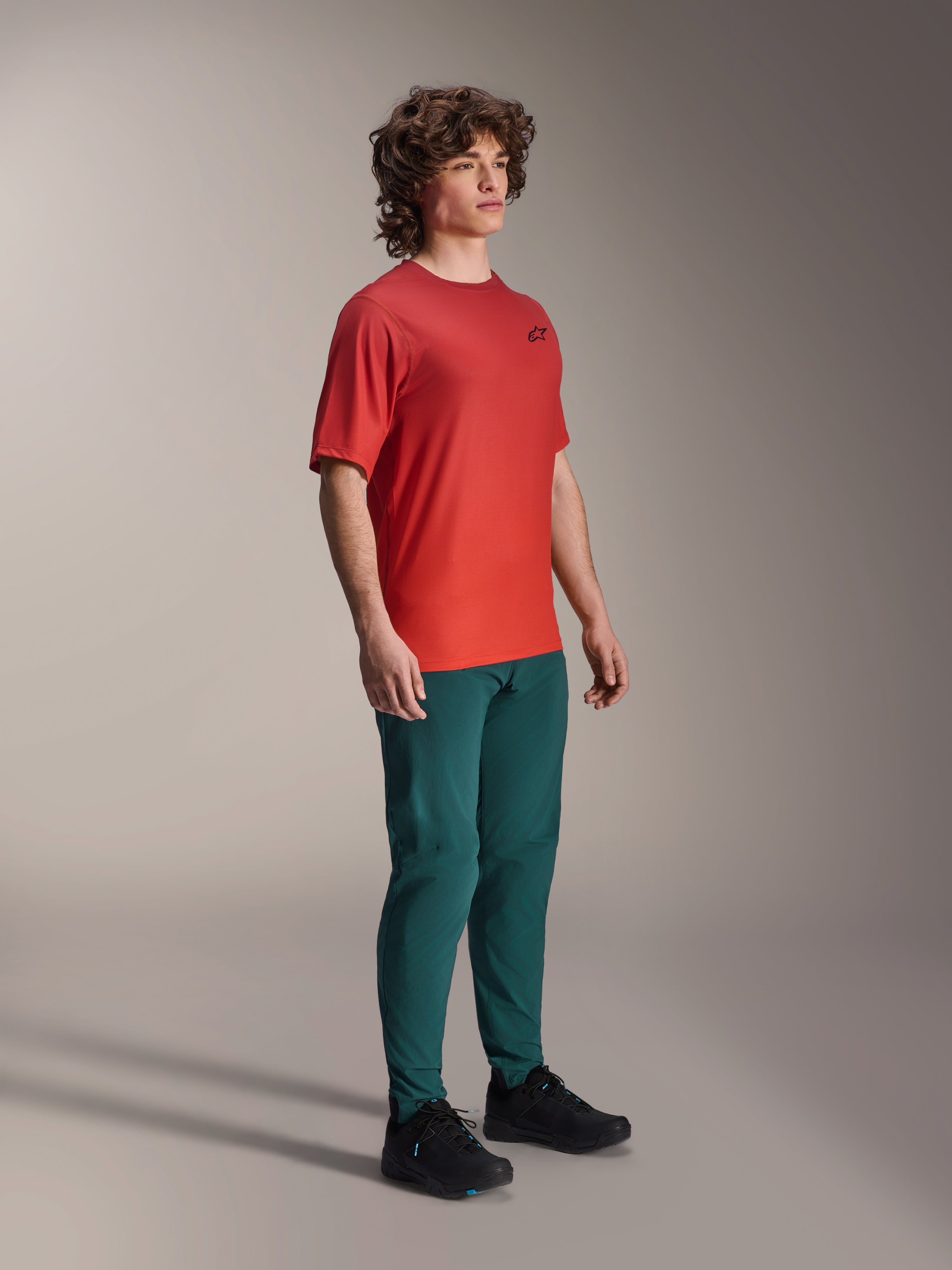 Modelo a usar calças Alpinestars A-Dura em Dark Pine, combinadas com uma camisola de manga curta A-Dura Rise vermelha e sapatos de BTT pretos, vestuário desportivo de mountain bike em ambiente de estúdio.