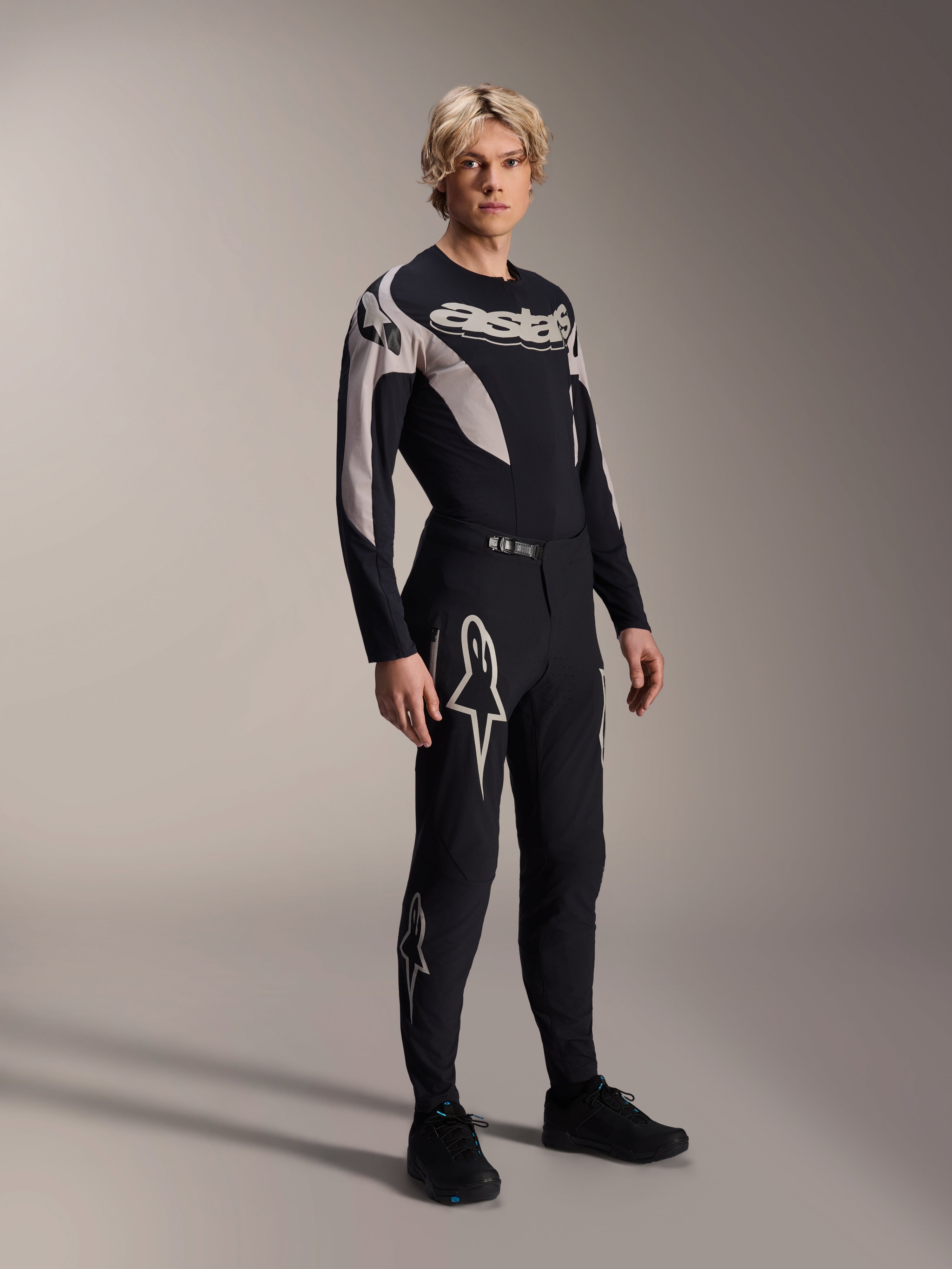 Modelo masculino a vestir a camisola Alpinestars A-Supra Melt - Manga Comprida, preta com pormenores gráficos cinzentos, logótipo 'astars' no peito, combinada com calças e sapatos pretos a condizer, kit de performance de mountain bike para corridas fora de estrada
