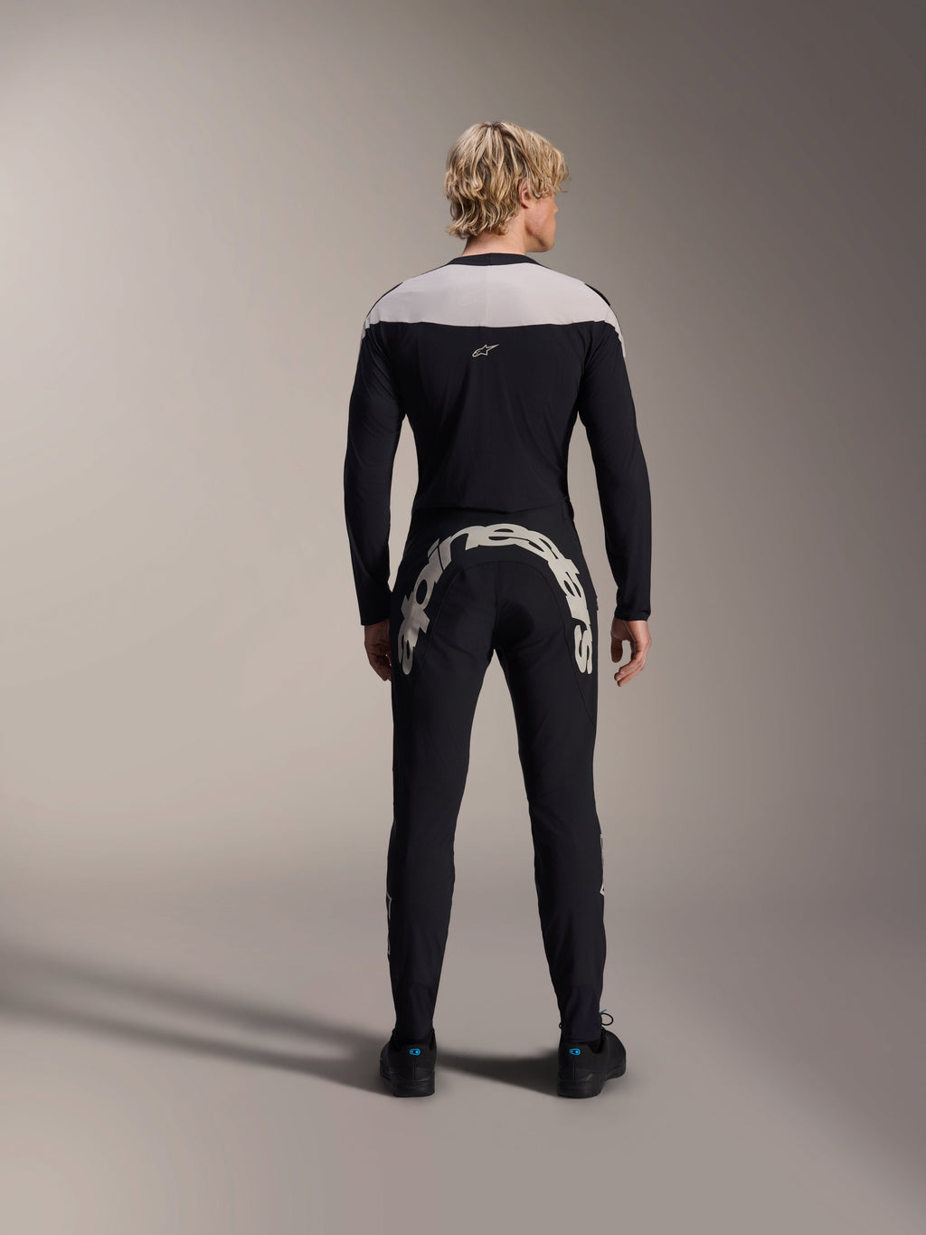 Calças Alpinestars A-Supra Melt, pretas, vista traseira a mostrar a grande marca nominativa Alpinestars branca na pala traseira, combinadas com uma camisola de manga comprida a condizer e sapatos de ciclismo pretos, equipamento de BTT de alto desempenho para condução todo-o-terreno.