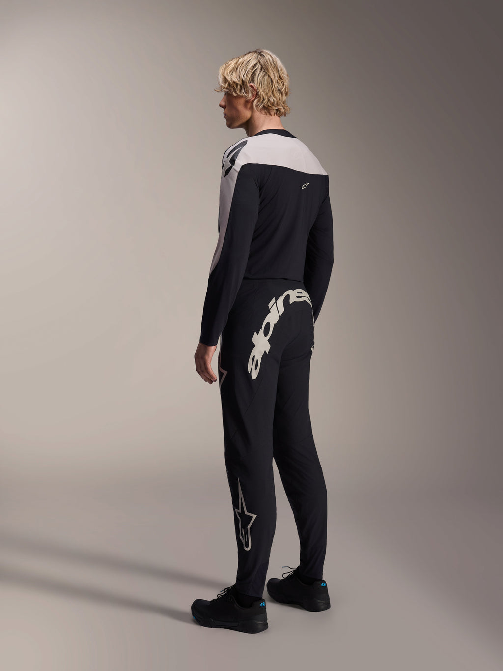 Calças Alpinestars A-Supra Melt, calças de MTB, pretas, com um grande logótipo Alpinestars branco na pala traseira e logótipo de estrela na perna, usadas por um modelo com uma camisola de manga comprida a condizer e sapatos pretos, vista lateral traseira