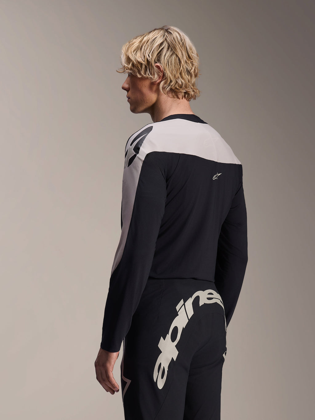 Modelo masculino a usar Camisola de Manga Comprida Alpinestars A-Supra Melt e Calças A-Supra Melt, equipamento de corrida de MTB preto e cinzento claro, com logótipo Alpinestars grande e branco em destaque na pala traseira das calças