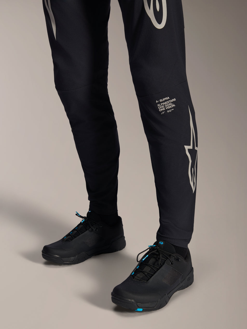 Calças Alpinestars A-Supra Melt, calças de BTT pretas, com a marca nominativa e o logótipo Alpinestars em branco na perna, combinadas com sapatos de ciclismo pretos com detalhes azuis, equipamento de performance para todo-o-terreno