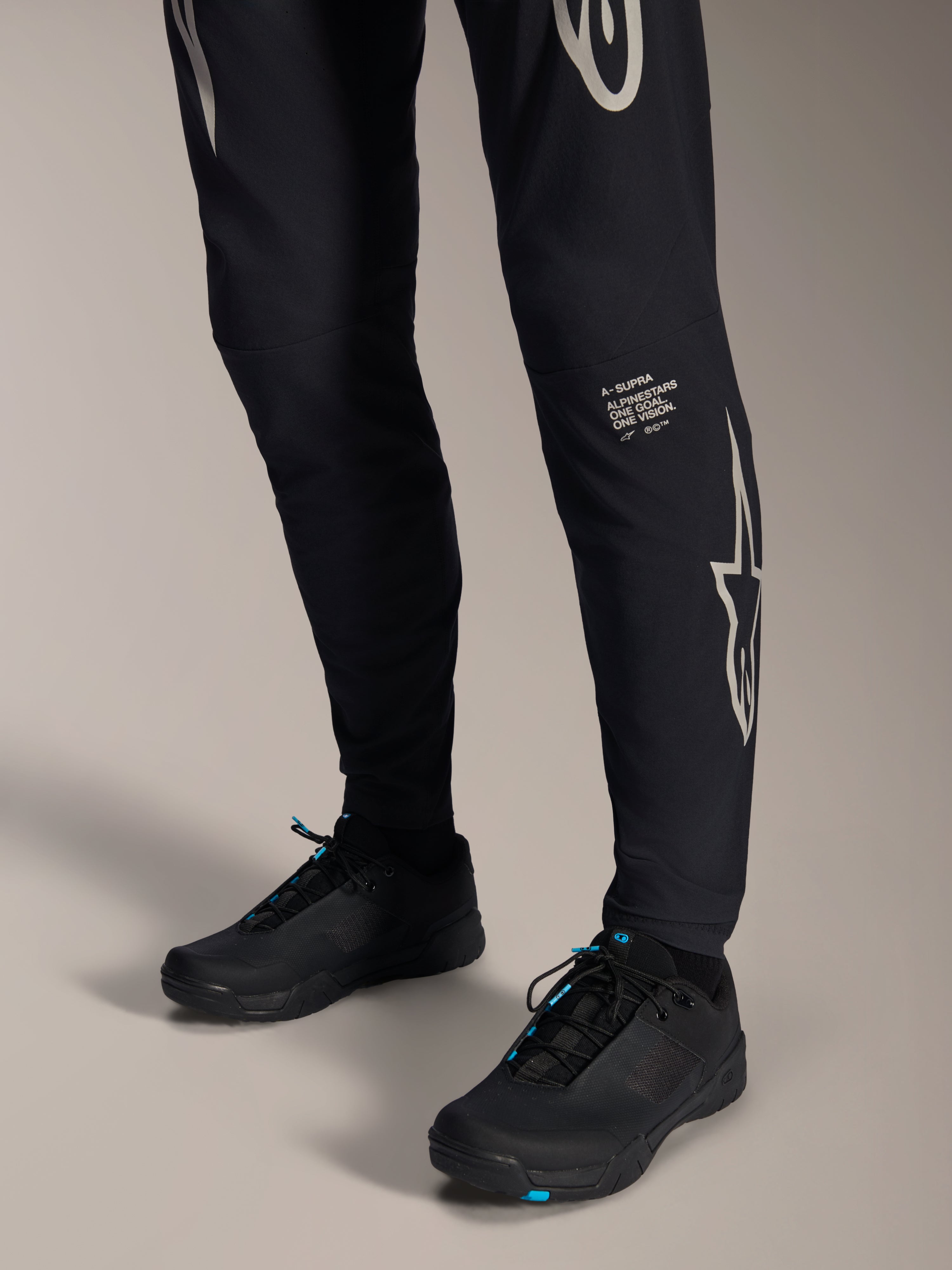 Calças Alpinestars A-Supra Melt, calças de BTT pretas, com a marca nominativa e o logótipo Alpinestars em branco na perna, combinadas com sapatos de ciclismo pretos com detalhes azuis, equipamento de performance para todo-o-terreno