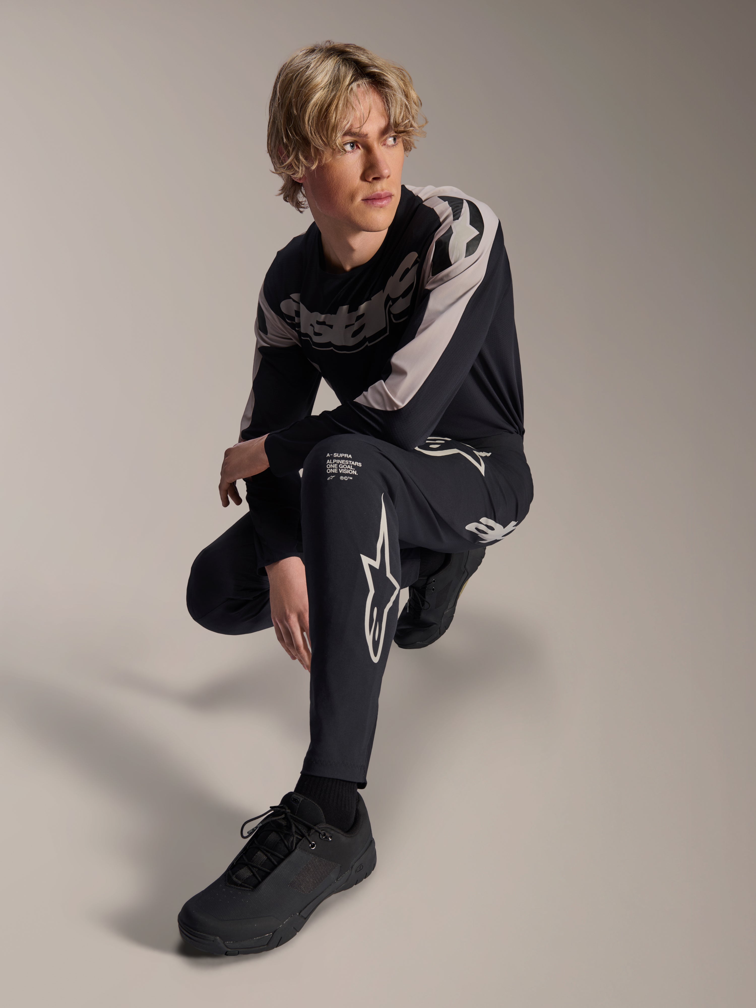 Calças e Jersey de Manga Comprida Alpinestars A-Supra Melt, pretas e cinzentas, com marca branca e logótipos de estrela em destaque, modelo masculino em pose agachada, equipamento de BTT coordenado