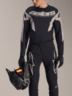 Kit de Corrida MTB Alpinestars A-Supra Melt, camisola de manga comprida e calças, preto e cinzento com logótipos Astars brancos, modelo a segurar num capacete integral preto e a usar luvas a condizer, equipamento de alta performance para BTT de downhill e enduro