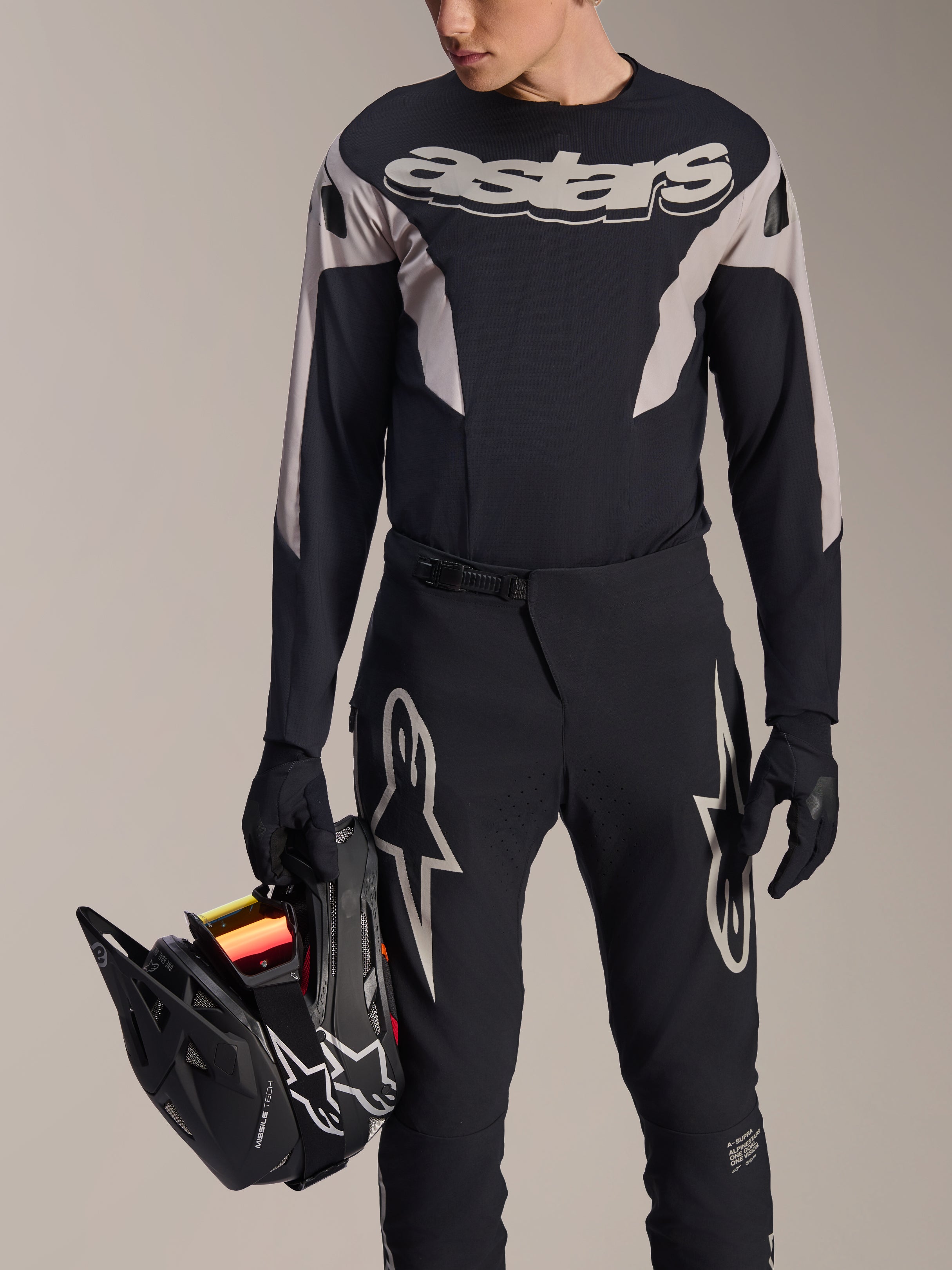 Kit de Corrida MTB Alpinestars A-Supra Melt, camisola de manga comprida e calças, preto e cinzento com logótipos Astars brancos, modelo a segurar num capacete integral preto e a usar luvas a condizer, equipamento de alta performance para BTT de downhill e enduro