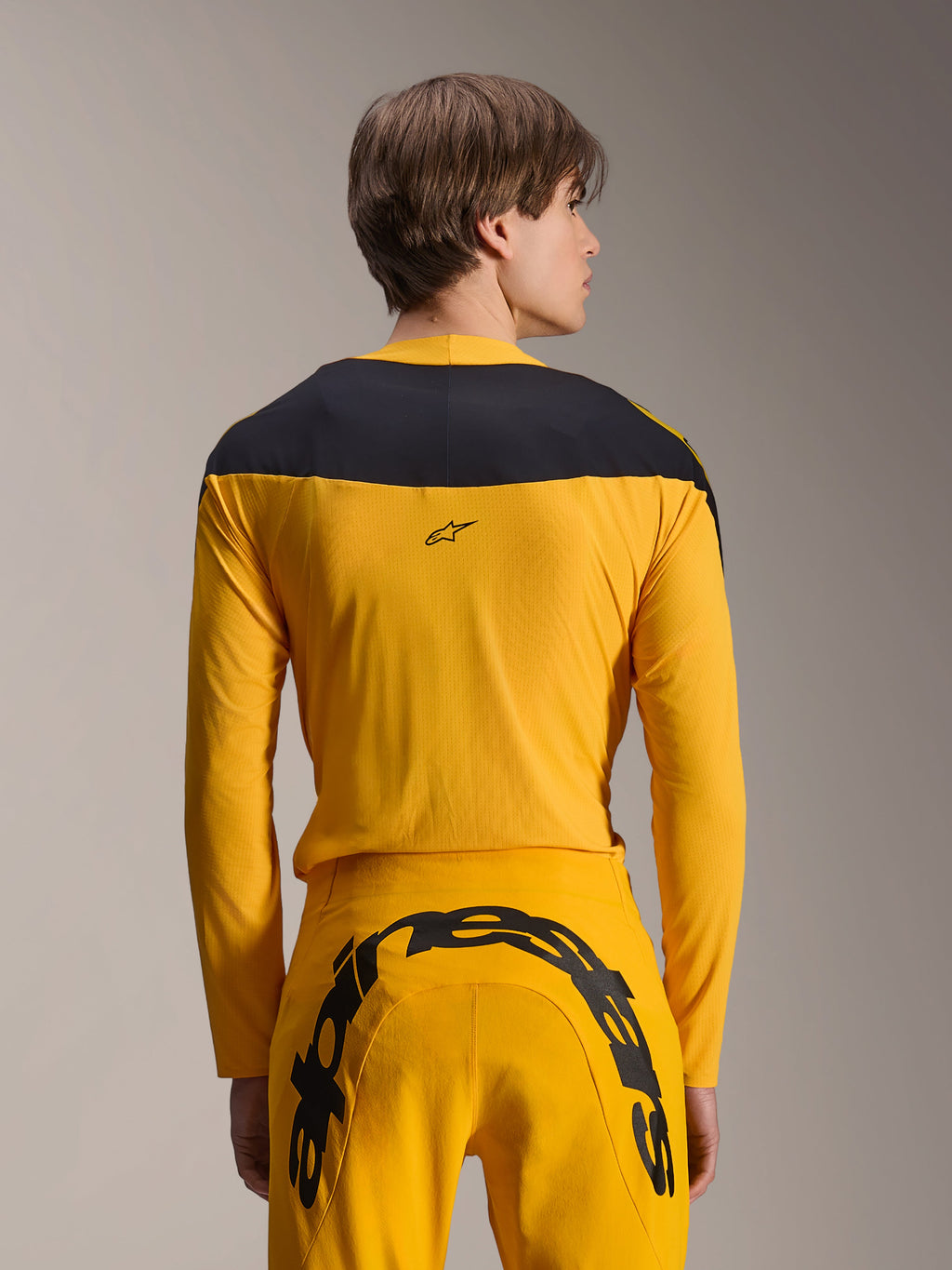 Alpinestars Limited Edition A-Supra Melt Gold Gear-Set, conjunto de equipamento de BTT amarelo, vista traseira de modelo a usar camisola de manga comprida com painel preto nos ombros e calças amarelas com um grande logótipo preto na parte traseira, vestuário de ciclismo de alta performance.