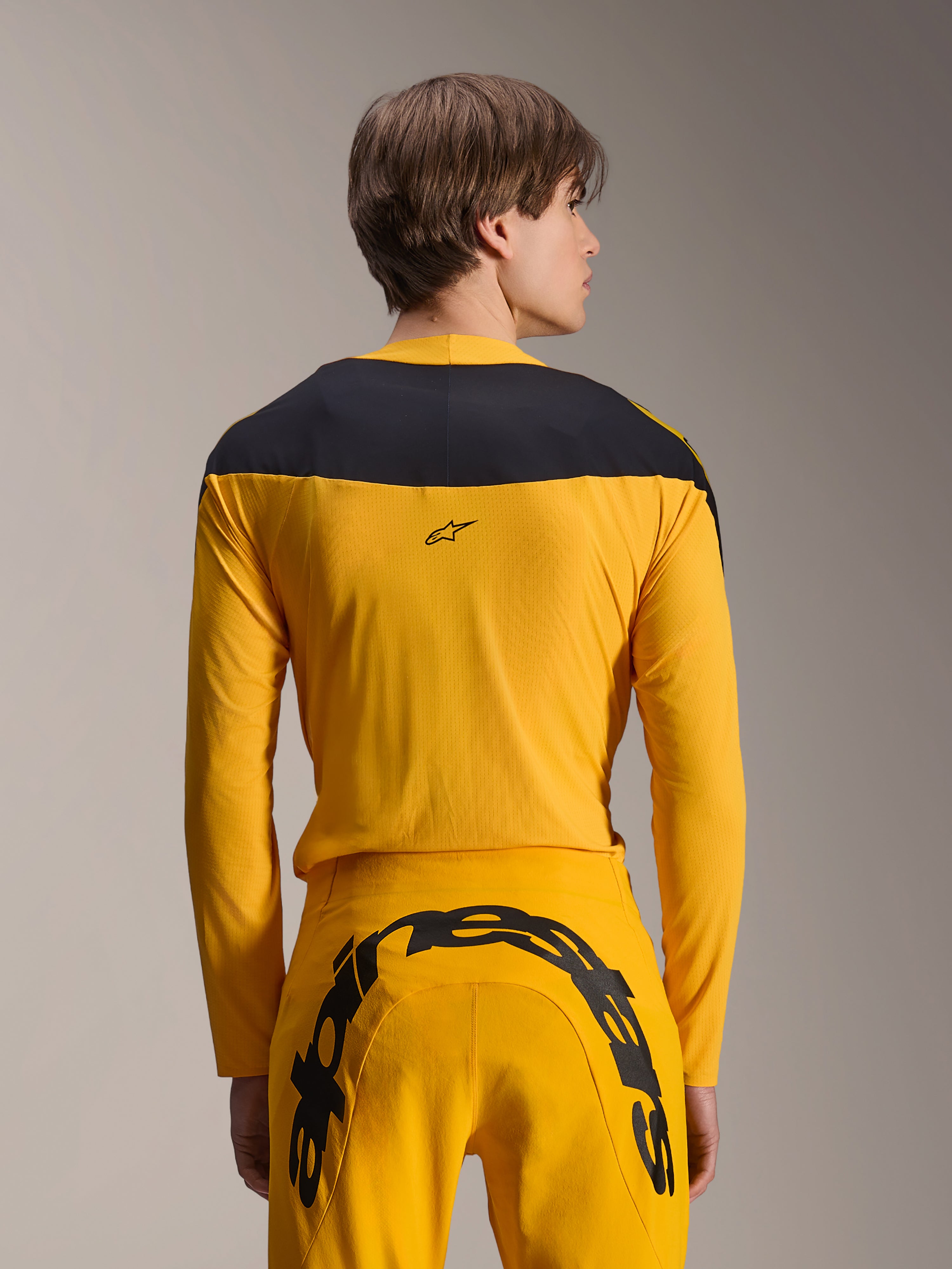 Alpinestars Limited Edition A-Supra Melt Gold Gear-Set, conjunto de equipamento de BTT amarelo, vista traseira de modelo a usar camisola de manga comprida com painel preto nos ombros e calças amarelas com um grande logótipo preto na parte traseira, vestuário de ciclismo de alta performance.
