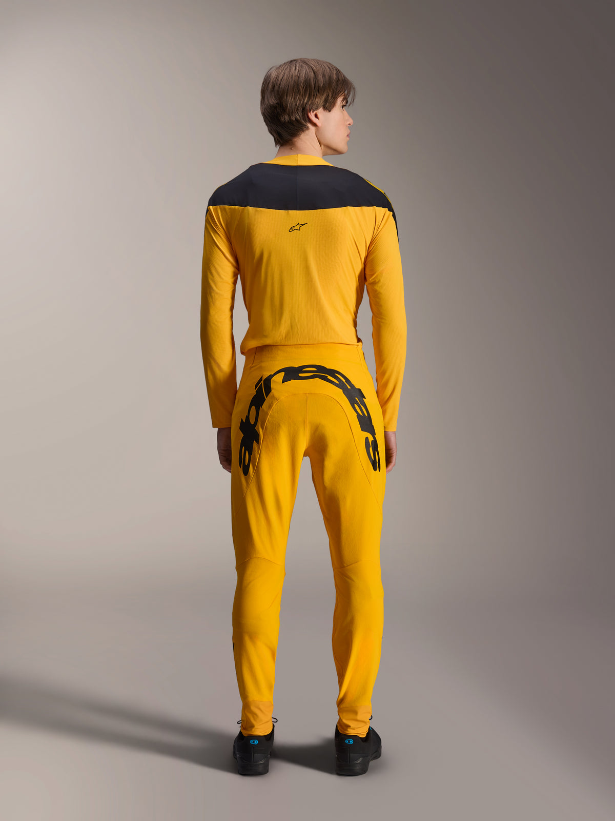Vista traseira de um modelo a usar o conjunto de equipamento Alpinestars Limited Edition A-Supra Melt Gold, com uma camisola de manga comprida amarelo escuro com encaixe preto nos ombros e calças amarelo escuro a condizer com um grande logótipo Alpinestars na pala traseira, combinado com sapatos pretos.
