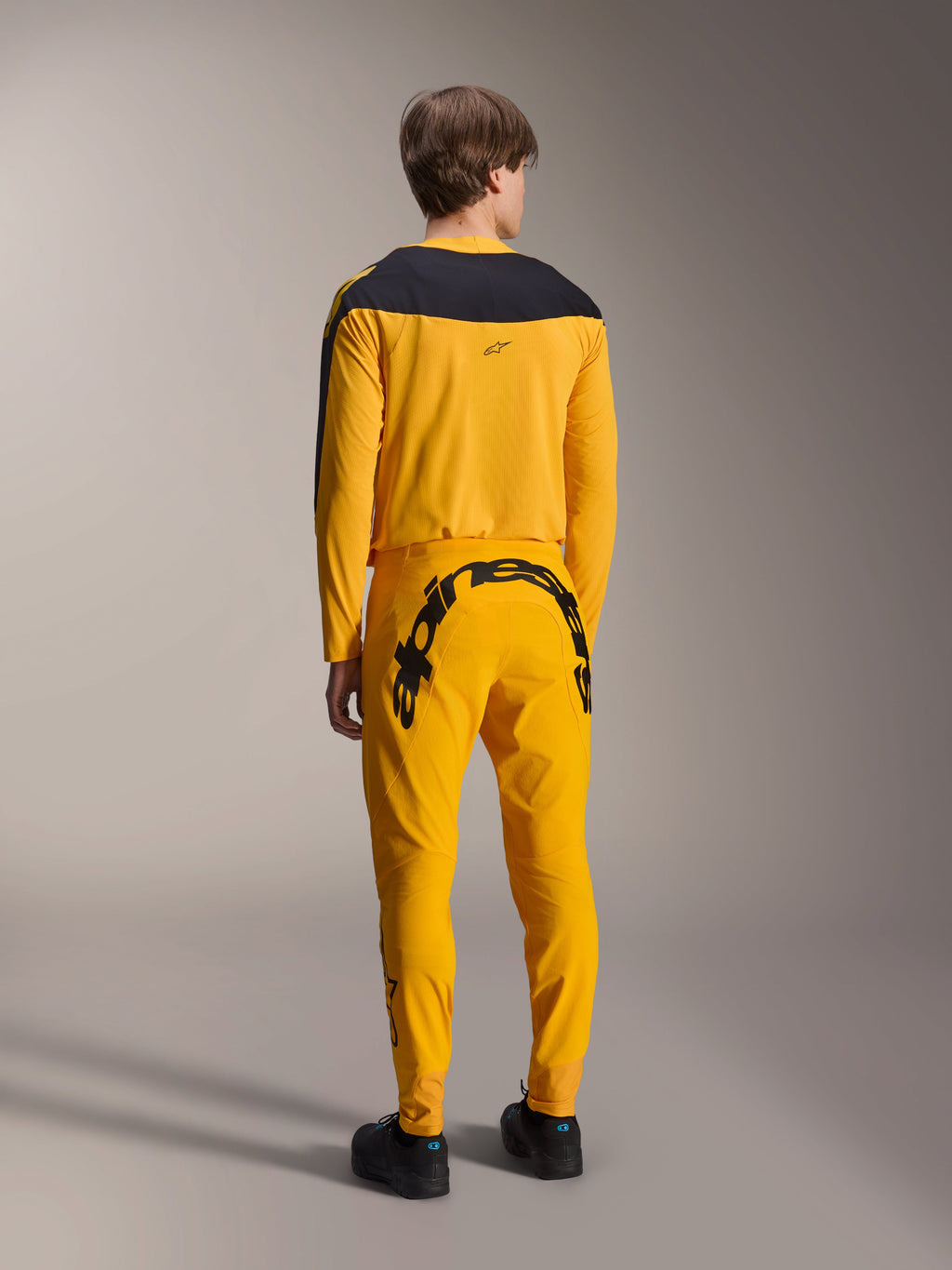 Camisola Alpinestars Limited Edition A-Supra Melt Gold, camisola de MTB de manga comprida, amarela escura com painel preto nos ombros, usada com calças amarelas a condizer com logótipo preto proeminente na parte de trás, vista traseira de conjunto de ciclismo de performance