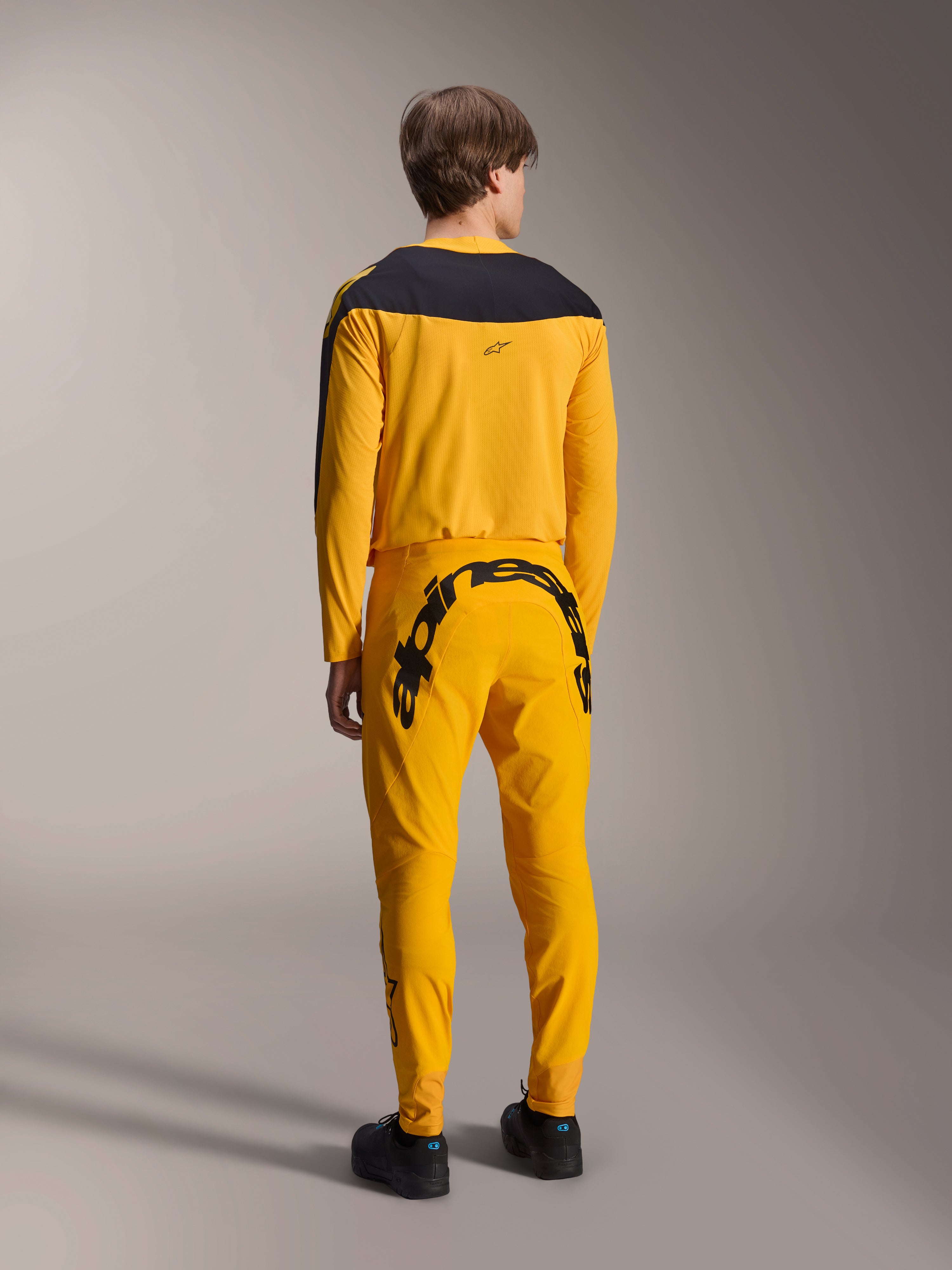 Camisola Alpinestars Limited Edition A-Supra Melt Gold, camisola de MTB de manga comprida, amarela escura com painel preto nos ombros, usada com calças amarelas a condizer com logótipo preto proeminente na parte de trás, vista traseira de conjunto de ciclismo de performance