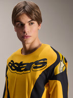 Camisola Alpinestars Limited Edition A-Supra Melt Gold, camisola de BTT de manga comprida, amarelo escuro com gráficos pretos inspirados em corridas, apresentando a inscrição 'astars' preta no peito e o logótipo de estrela no ombro, vestida por um modelo masculino