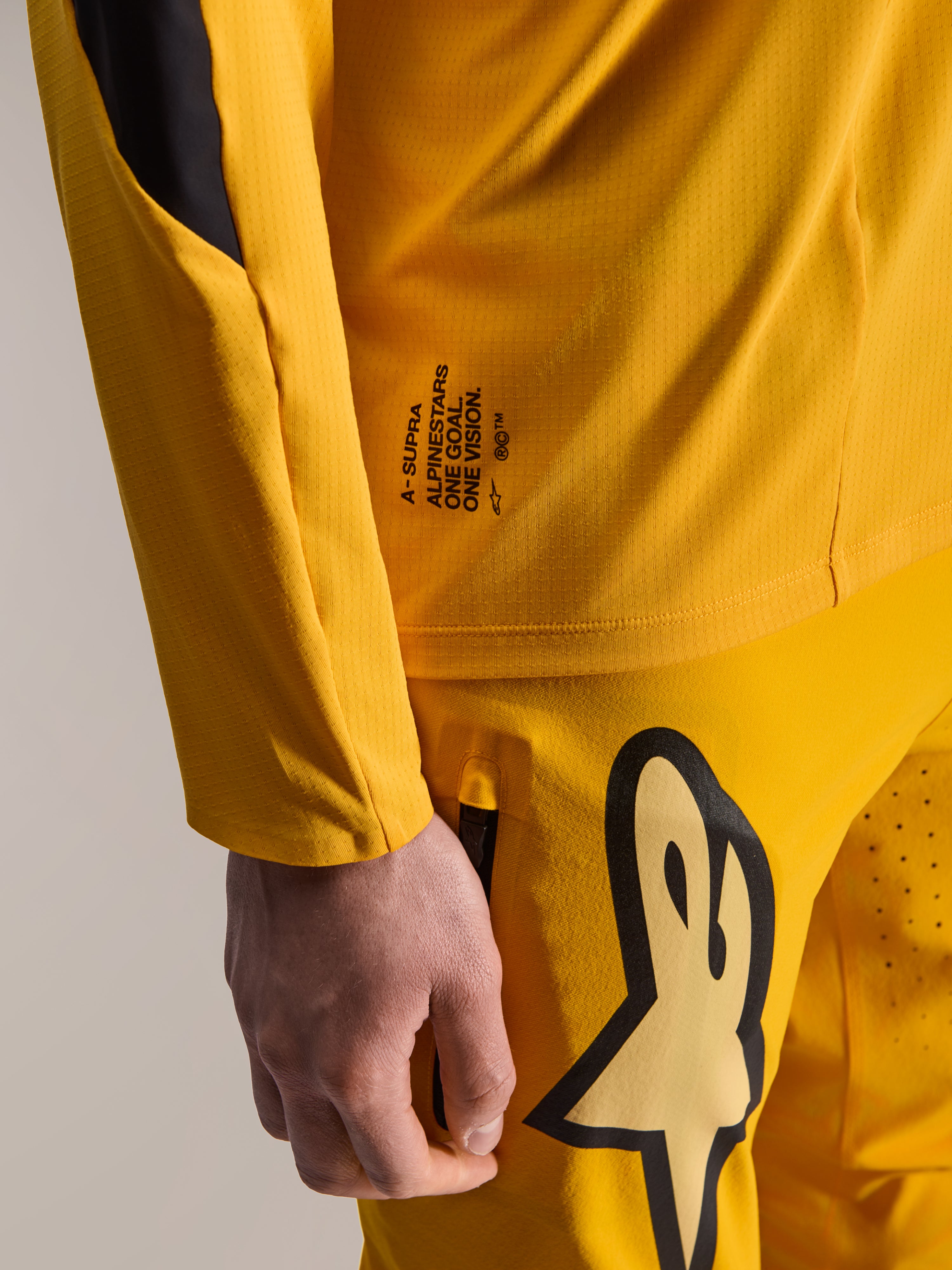 Calças Alpinestars Limited Edition A-Supra Melt Gold e camisola de manga comprida a condizer, amarelo escuro, vista em pormenor com a marca 'One Goal. One Vision.' e logótipo estilizado grande na coxa, equipamento de BTT de alta performance