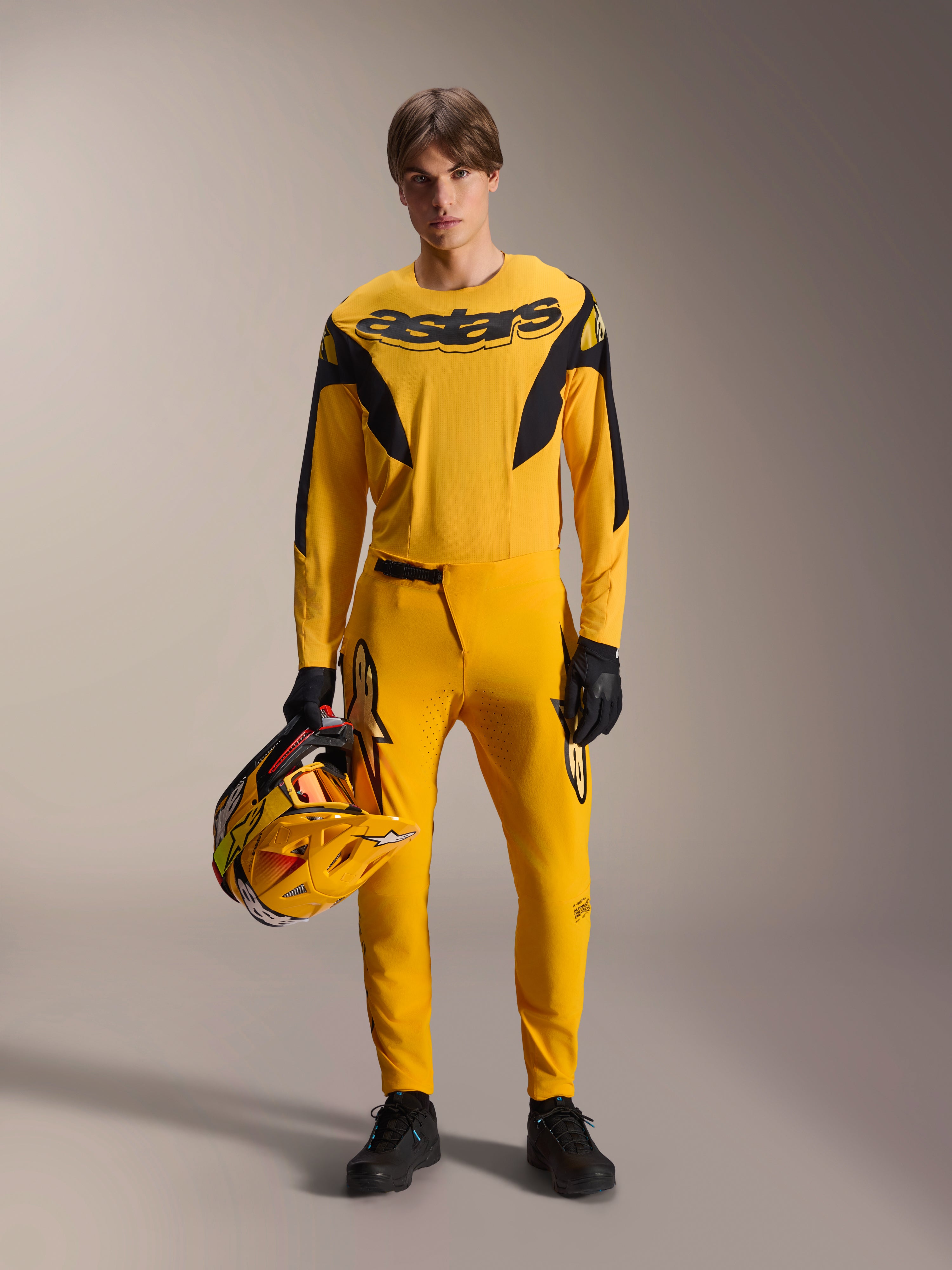 Modelo a usar o Kit de MTB Alpinestars Limited Edition A-Supra Melt Gold, camisola amarela de manga comprida com logótipo 'astars' preto, calças amarelas a condizer com detalhes pretos, segurando um capacete de corrida amarelo e preto, luvas e sapatos pretos, equipamento de performance para BTT